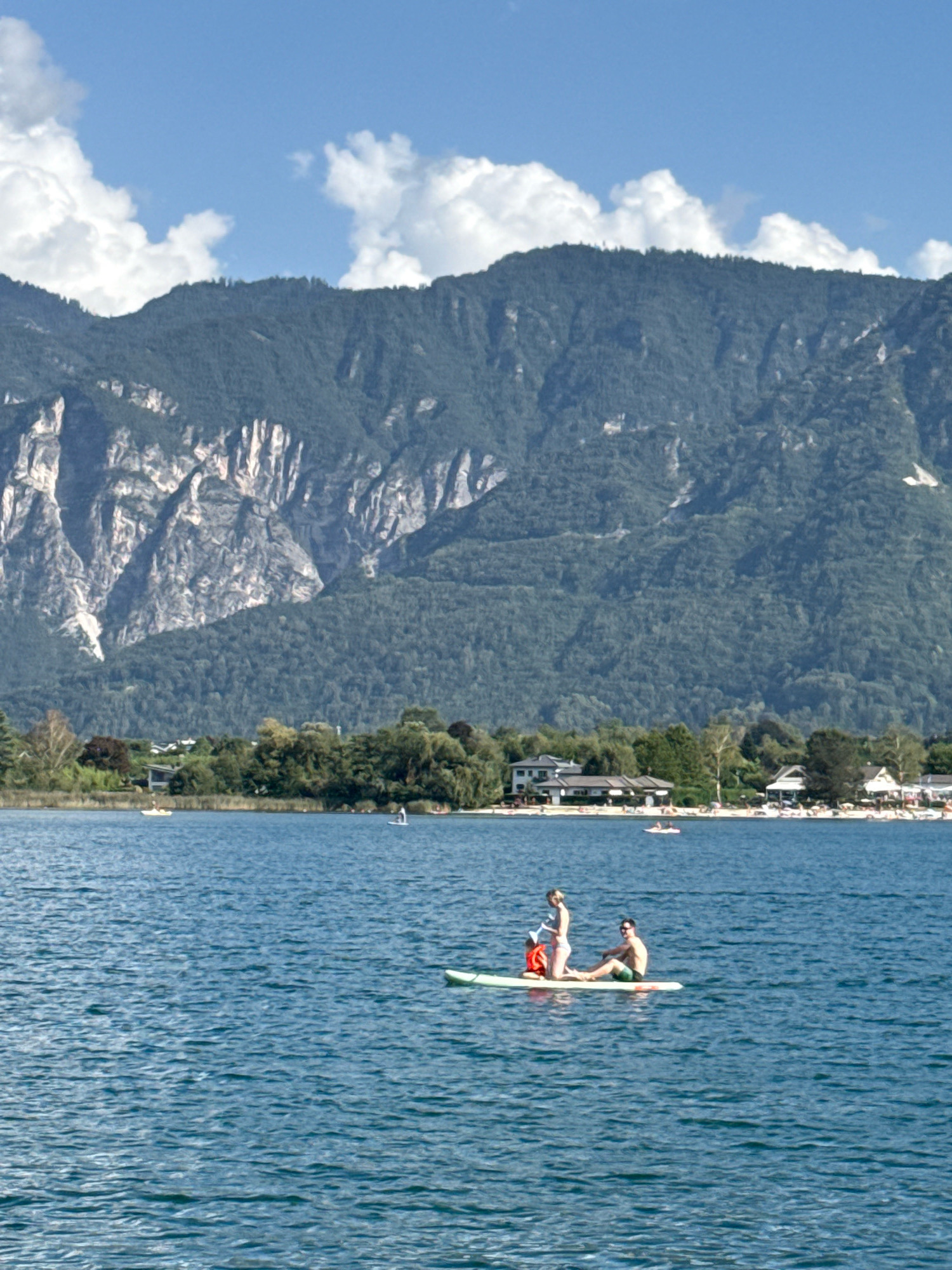 The image captures a serene scene on a body of water with a mountainous backdrop. In the foreground, a paddleboard with two individuals is visible. A woman is kneeling on the paddleboard, and a man is sitting behind her. They are both looking forward. The paddleboard is a light green color. The water is a deep blue with ripples on the surface, suggesting a gentle breeze.
In the middle ground, the shore of a town is lined with trees and buildings. Several people are visible on the shore, some appearing to be sunbathing on what looks like a beach. Other small boats and individuals on paddleboards are scattered across the water further out. The buildings are modern in style with white and beige facades and dark roofs.
The background is dominated by a large, verdant mountain range. The slopes of the mountain are covered in dense green foliage, with visible rock formations and crevices on the steeper sections. The sky above is a clear blue with scattered white clouds. The overall impression is one of a recreational day in a picturesque natural setting, likely during warmer weather. The location is Calceranica al Lago, Italy.