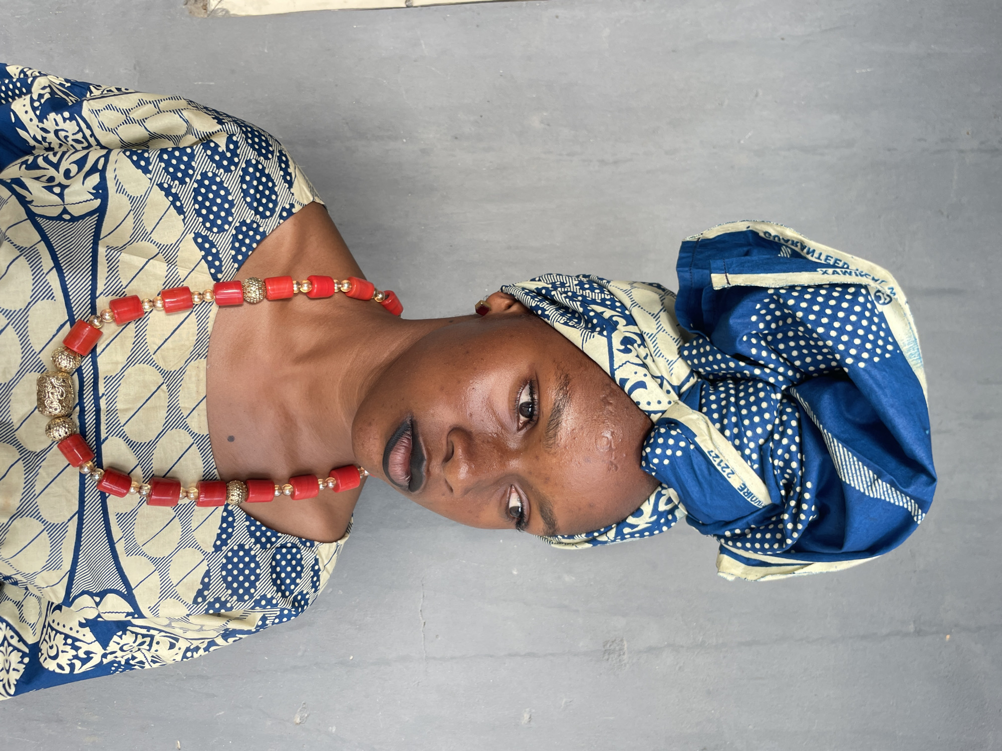 A woman is centered in the frame, wearing a traditional blue and cream patterned dress and a matching headwrap. She has a red beaded necklace around her neck. The background appears to be a gray, possibly concrete, surface. The image has been rotated slightly. The location is Badagry, Nigeria.