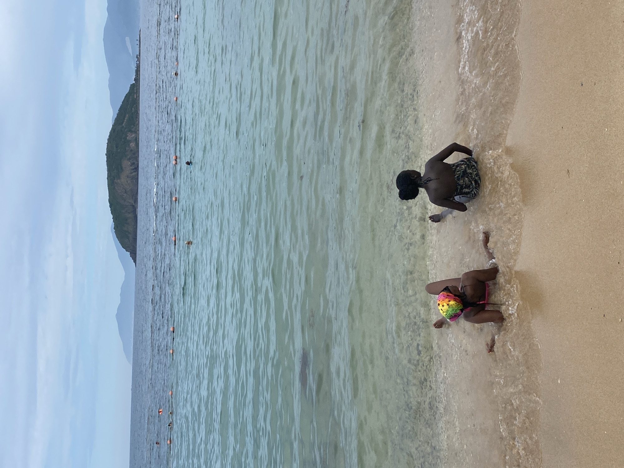 Two young women are sitting by the shore of a beach in Nha Trang, Vietnam. The woman on the left is lying on her side, facing away from the camera, with her head propped up on her hand. She is wearing a colorful, patterned headscarf and a pink bikini. The woman on the right is sitting with her back to the camera, facing the ocean. She is wearing a black and white patterned swimsuit. Both women are partially submerged in the shallow water. The sand is a light tan color, and the water is clear and turquoise. In the distance, there are several buoys floating in the water, and a small island with a green, tree-covered hill can be seen. The sky is overcast with a few wispy clouds.