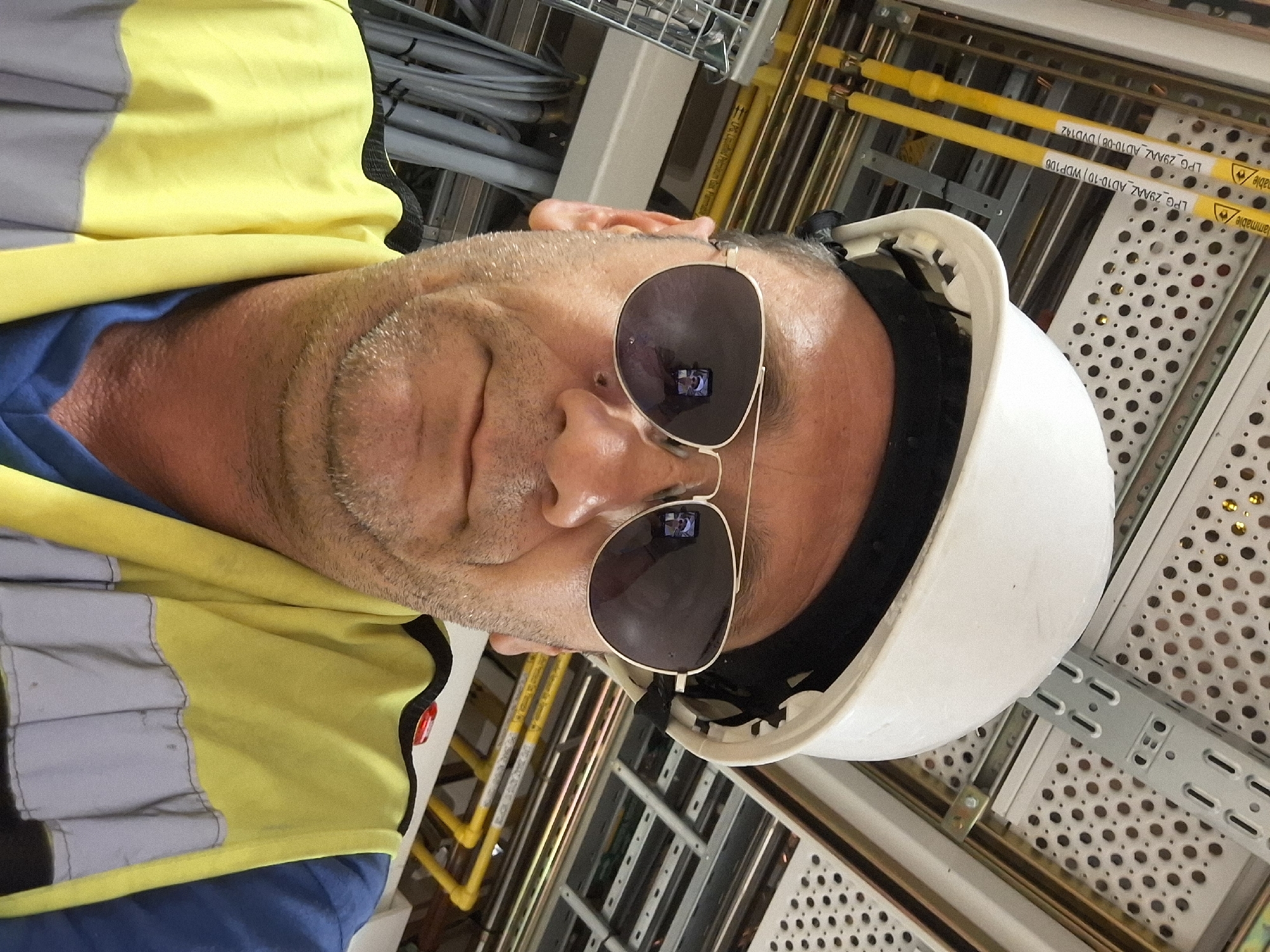 A man wearing a white hard hat, aviator sunglasses, and a yellow and gray high-visibility vest is taking a selfie. He is inside a building with exposed wiring, conduits, and metal framing on the ceiling, characteristic of an industrial or construction site. The background shows yellow pipes labeled "LPG 29MAZ AD10-08 DV0142" and "LPG 29MAZ AD10-10 WDP1DB", indicating a facility related to Liquefied Petroleum Gas. The man has stubble and is looking directly at the camera. The image appears to be taken at a facility in Kiryat Gat, Israel, as suggested by the context of its origin.