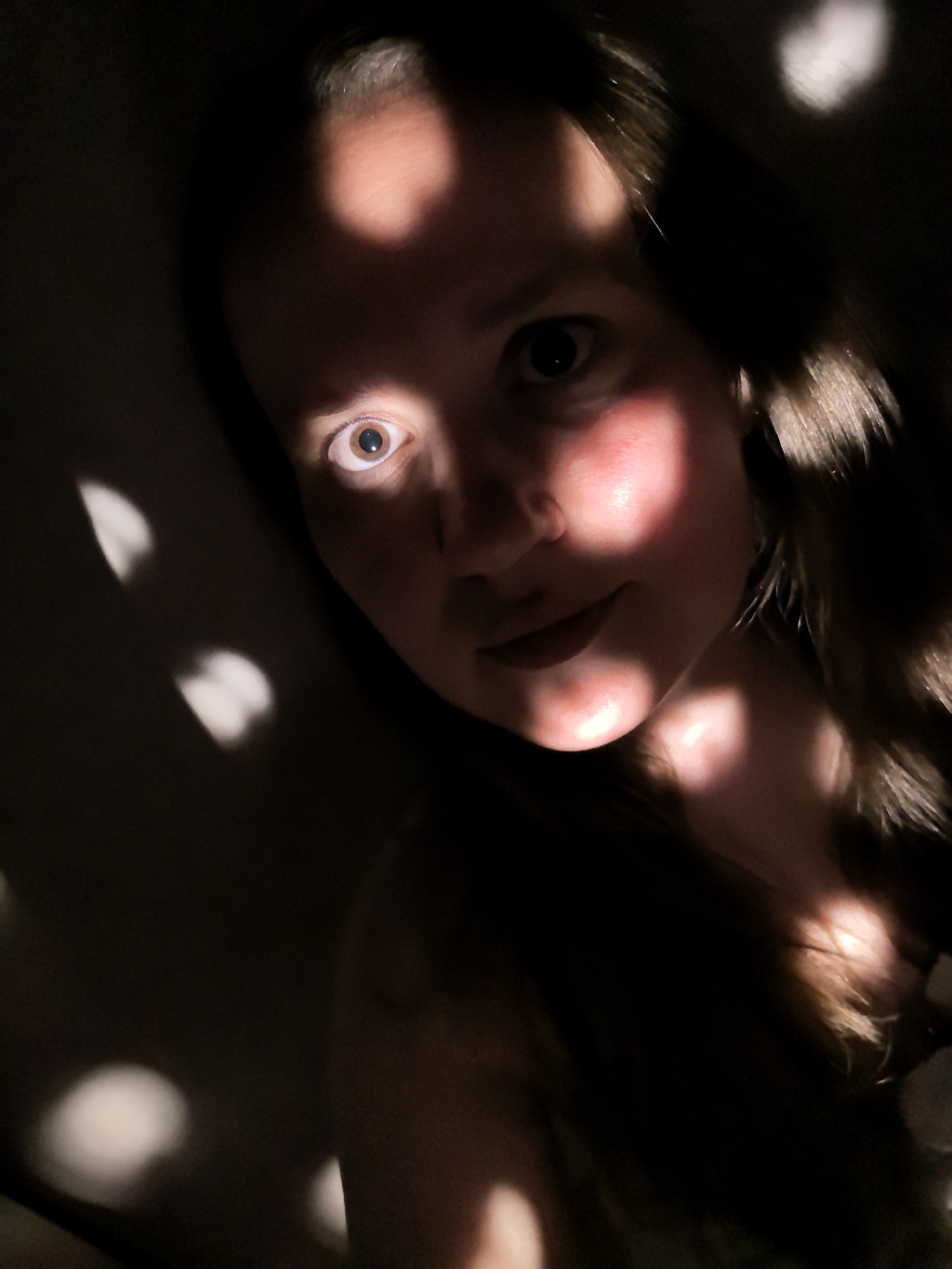 A female human with light skin is the primary subject in this darkly lit indoor photograph. Her head is tilted slightly, and her gaze is directed toward the camera. The right side of her face is illuminated, while the left side is cast in shadow. A pattern of diffuse, circular highlights falls across the wall behind her and on her face, possibly caused by light filtering through foliage or a patterned screen. The photograph is undated and contains no overt indication of being located in the city of Фролы, Russia. The context of this image is limited.