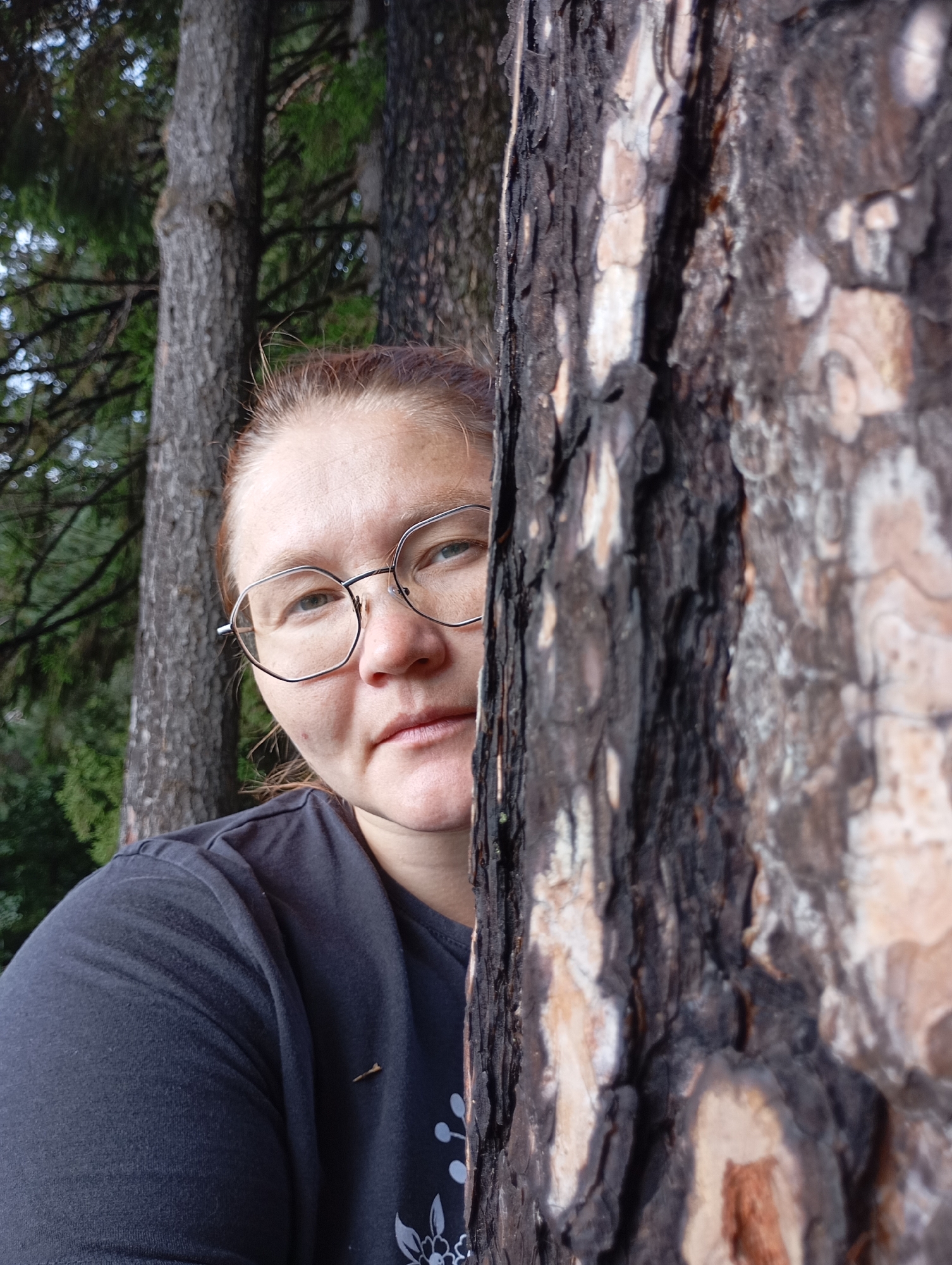 The image shows a woman with reddish-brown hair and glasses peeking from behind a tree trunk. She is wearing a dark t-shirt. The background consists of trees and foliage, suggesting an outdoor setting. The image does not provide any specific details about the city of Chaikovsky, Russia, or any other identifiable location. The scene appears to be a candid portrait taken in a natural environment.