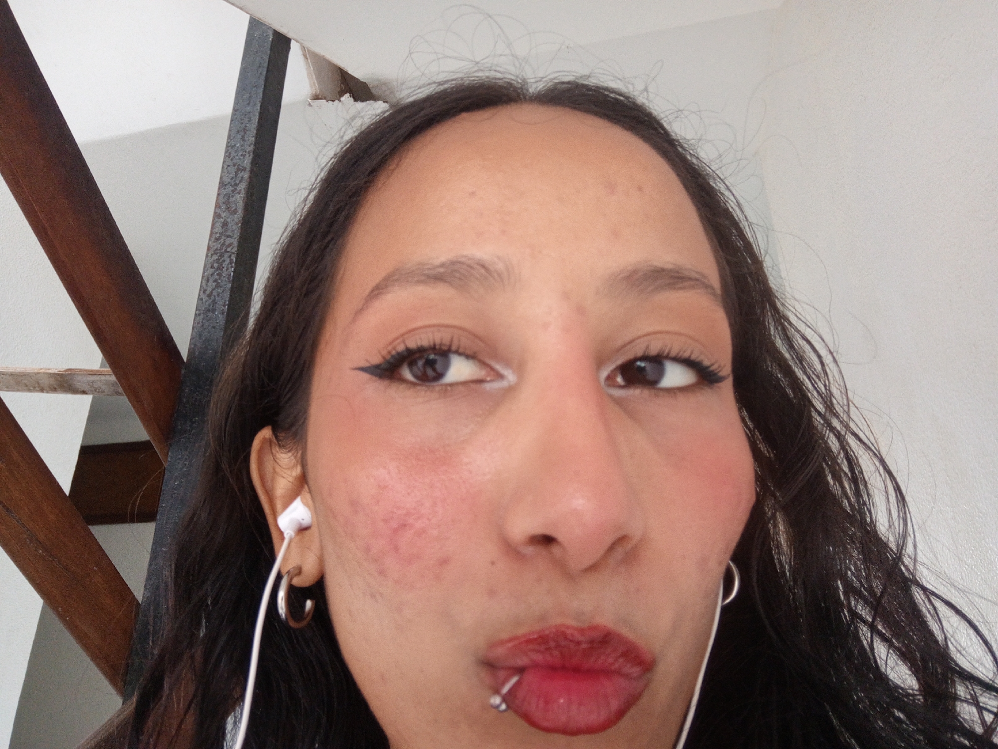 A close-up selfie depicts a young woman with dark hair, light brown skin, and dark eyes. Her facial features include a prominent nose and full lips, adorned with a lip piercing on the lower lip. She is wearing dark eyeliner, white eyeshadow, and red lipstick. She also wears silver earrings. She is wearing white headphones.
The background shows a staircase and a white wall. The location cannot be accurately determined to be in Porlamar, Venezuela, based on the image.