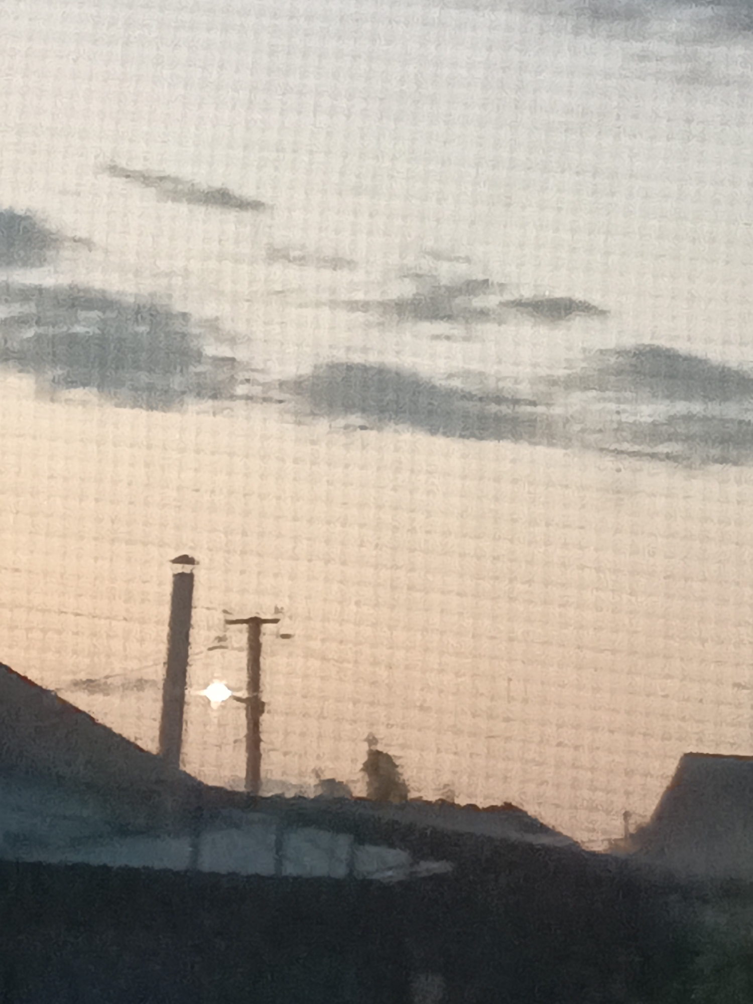 The image captures a hazy, overcast sky with scattered clouds, tinged with the pale hues of sunrise or sunset. Silhouetted against this backdrop are the dark shapes of rooftops, suggesting an urban or suburban environment. A prominent utility pole with electrical wires stands near the center, and beside it, a vertical structure, possibly a chimney or another pole, rises from a rooftop. The sun is partially obscured by the pole and a cloud, creating a bright, diffused glow. The foreground is dominated by dark, indistinct shapes, likely more buildings or structures. The overall impression is one of a quiet, possibly early morning or late evening scene in the city of Tbilisskaya, Russia.