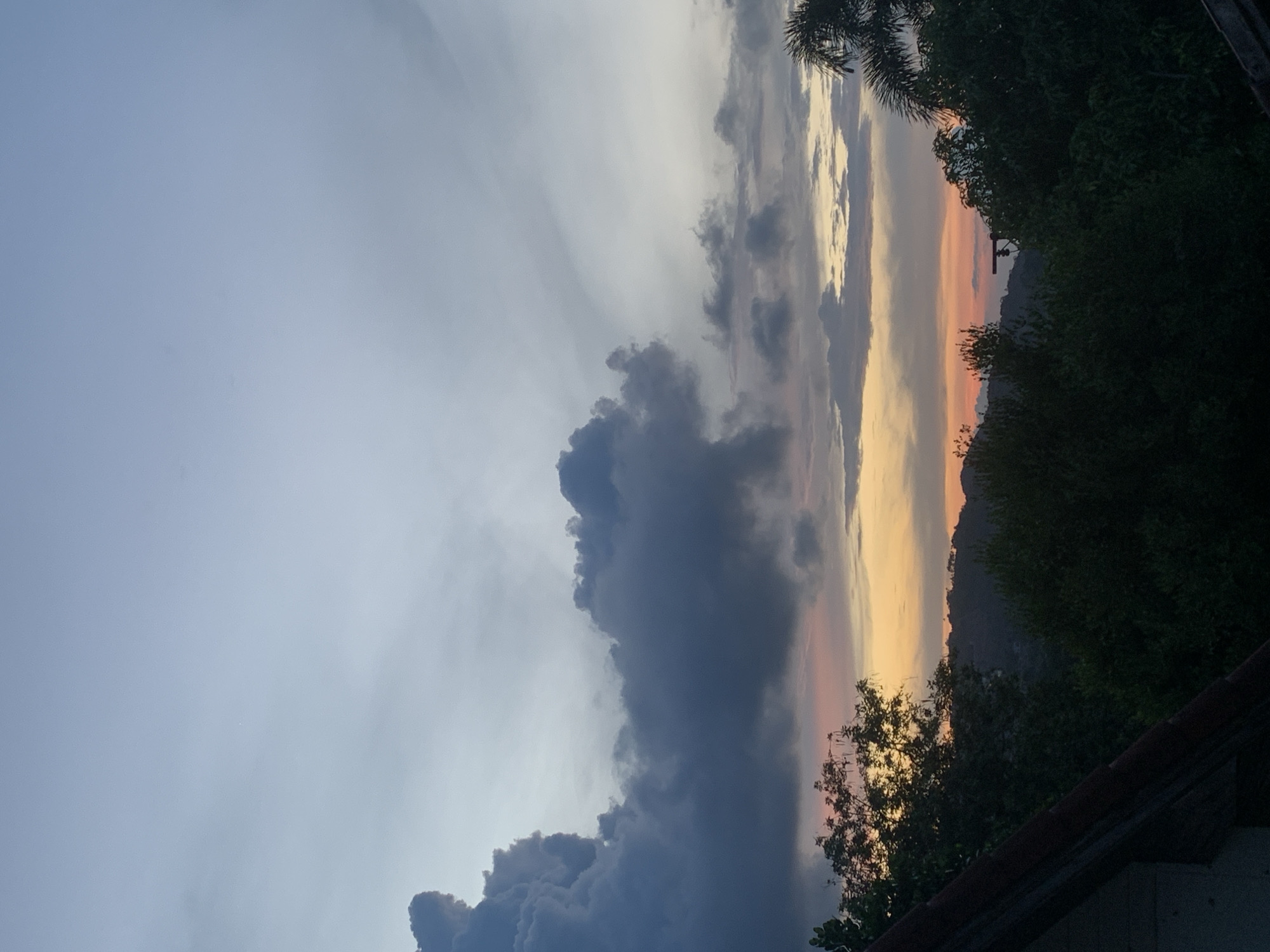 The image shows a dusk or dawn scene in บ้านบางรักษ์, Thailand. The sky is predominantly filled with clouds of varying shades, from light gray to dark blue-gray. A vertical band of orange and pink light indicates the rising or setting sun. Lush greenery and trees obscure the distant horizon and a mountain ridge. A portion of a roof is visible in the foreground. A utility pole or electrical wire is silhouetted against the colorful sky.