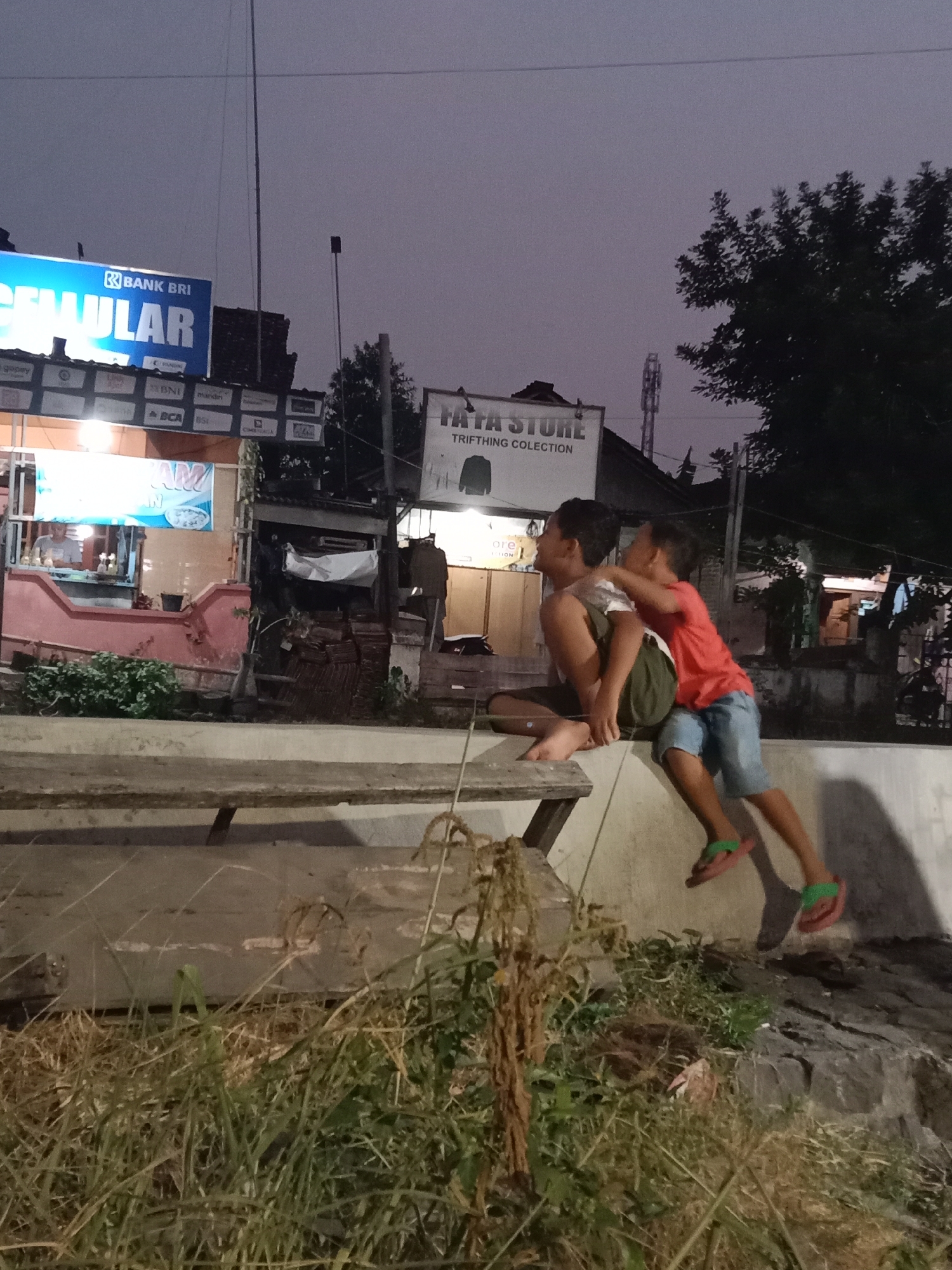 The image captures an outdoor scene at dusk, likely in Langensari, Indonesia, with two young boys interacting. One boy, wearing a red t-shirt and blue shorts, is playfully embracing another boy who is sitting down. The seated boy wears a dark green t-shirt and olive shorts. They are positioned on a concrete ledge or wall, with a wooden bench-like structure to their left. In the background, commercial signage is visible, including a prominent blue sign that reads "BANK BRI" and "SELULAR", and another sign that says "TATA STORE" and "TRIFTHING COLLECTION". A small food stall with a person inside is also visible to the left, with bright lighting emanating from it. The environment appears to be a roadside or a public area, with vegetation and some debris present in the foreground. The overall lighting suggests it is early evening.