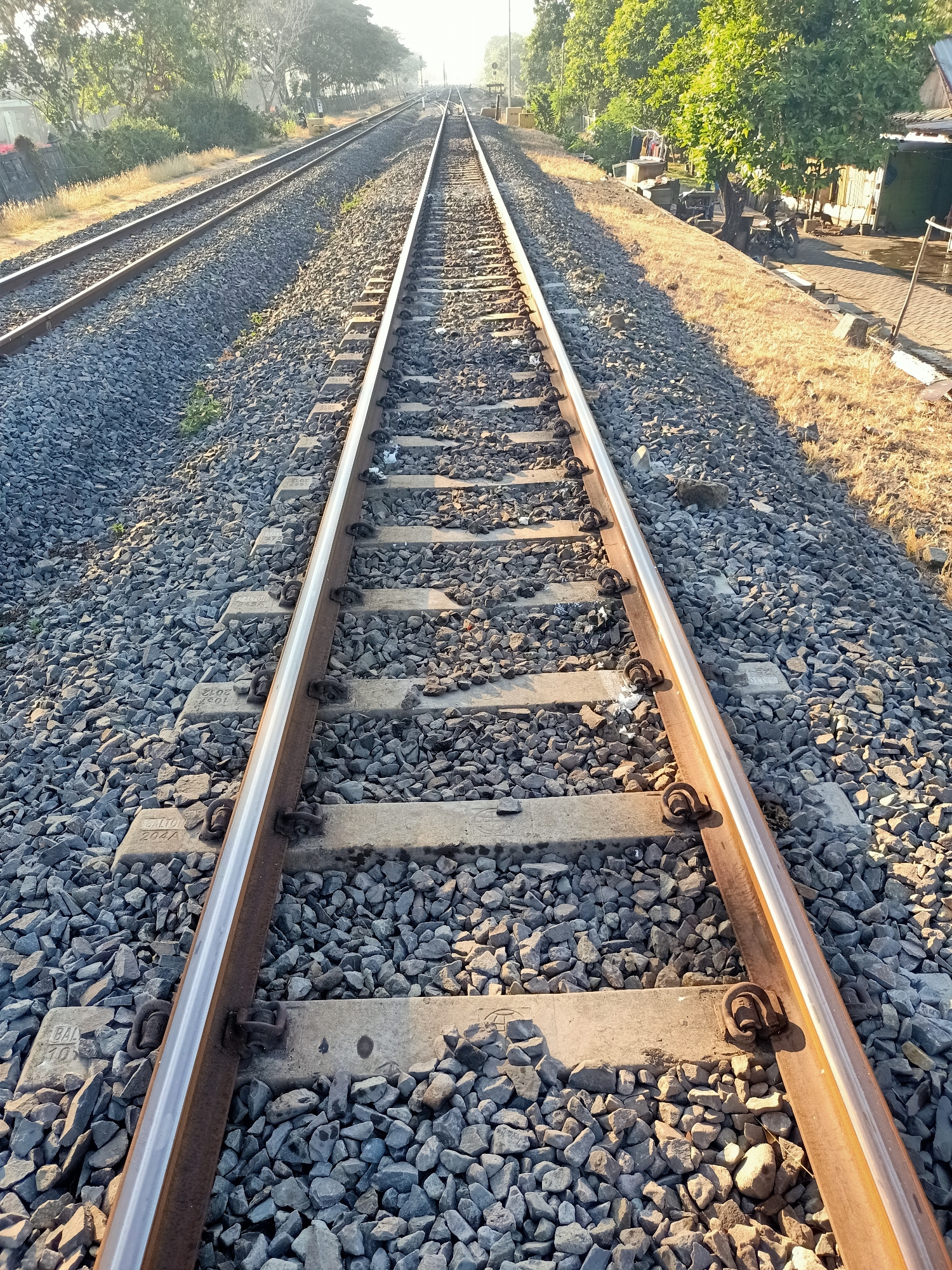 The image depicts a railway track in Kendung, Indonesia, under a bright, overcast sky. The perspective is from ground level, looking down the parallel tracks. The tracks are made of steel rails secured to concrete sleepers laid on a bed of grey gravel. The gravel is interspersed with sparse green vegetation. To the right of the tracks, there's a raised embankment covered in dry, yellow grass, with trees lining the top. A small building with a corrugated metal roof and some parked motorcycles are visible on this embankment, suggesting a rural or semi-urban setting. The left side of the image shows another set of tracks running parallel, disappearing into the distance. The overall atmosphere is one of stillness and early morning light, with long shadows cast across the tracks.
