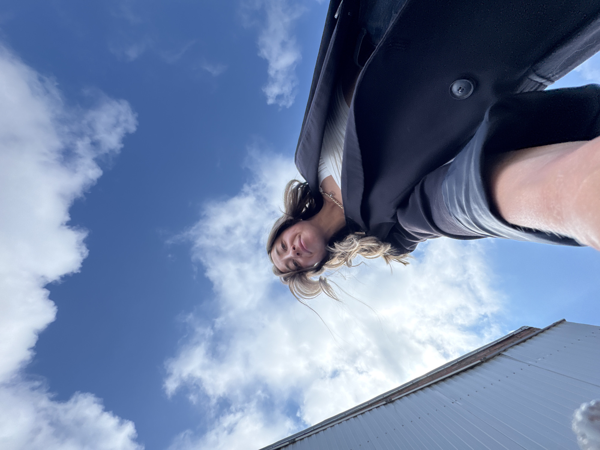 This outdoor shot, taken from a low-angle perspective looking upwards, features a woman against a bright blue sky with scattered white clouds. The woman's face is visible in the upper central part of the frame, looking down towards the camera with a slight smile. She has light-colored hair, some strands appearing to move in a breeze. She is wearing a dark coat or jacket over a light-colored ribbed top, and part of her arm and hand are visible on the right. The dominant background is a clear, vibrant blue sky punctuated by fluffy white clouds. In the bottom right corner, the top edge of a light-colored building, possibly an industrial structure or warehouse with a panelled roof, is visible. The scene is brightly lit, indicating it is daytime. The overall impression is casual and pleasant. This image is set in Chaikovsky, Russia. No visible text is present.