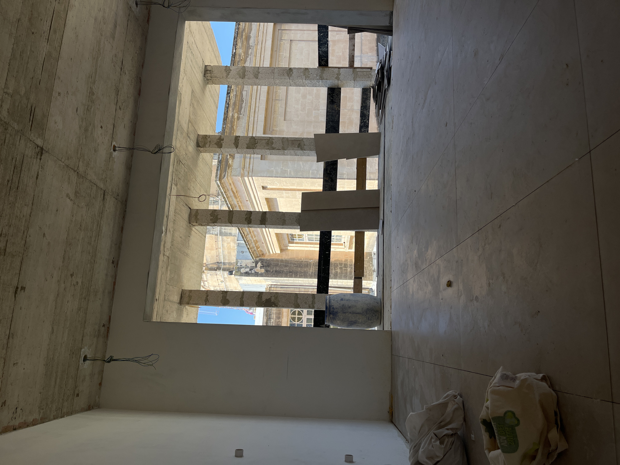 The image shows an interior view of an unfinished building under construction, likely an apartment or office space. The walls are bare concrete or plaster, and the ceiling is also exposed concrete, with electrical wires hanging down. The floor is tiled with large, pale tiles. A large opening in the wall looks out onto a street scene, with a building of classical architecture featuring columns and arched windows visible in the distance. There is a blue barrel and some construction materials, including large panels and stacks of what appear to be smaller tiles or planks, near the opening. A few bundles of fabric or sheeting are on the floor in the foreground. The scene is well-lit by natural light coming through the opening.