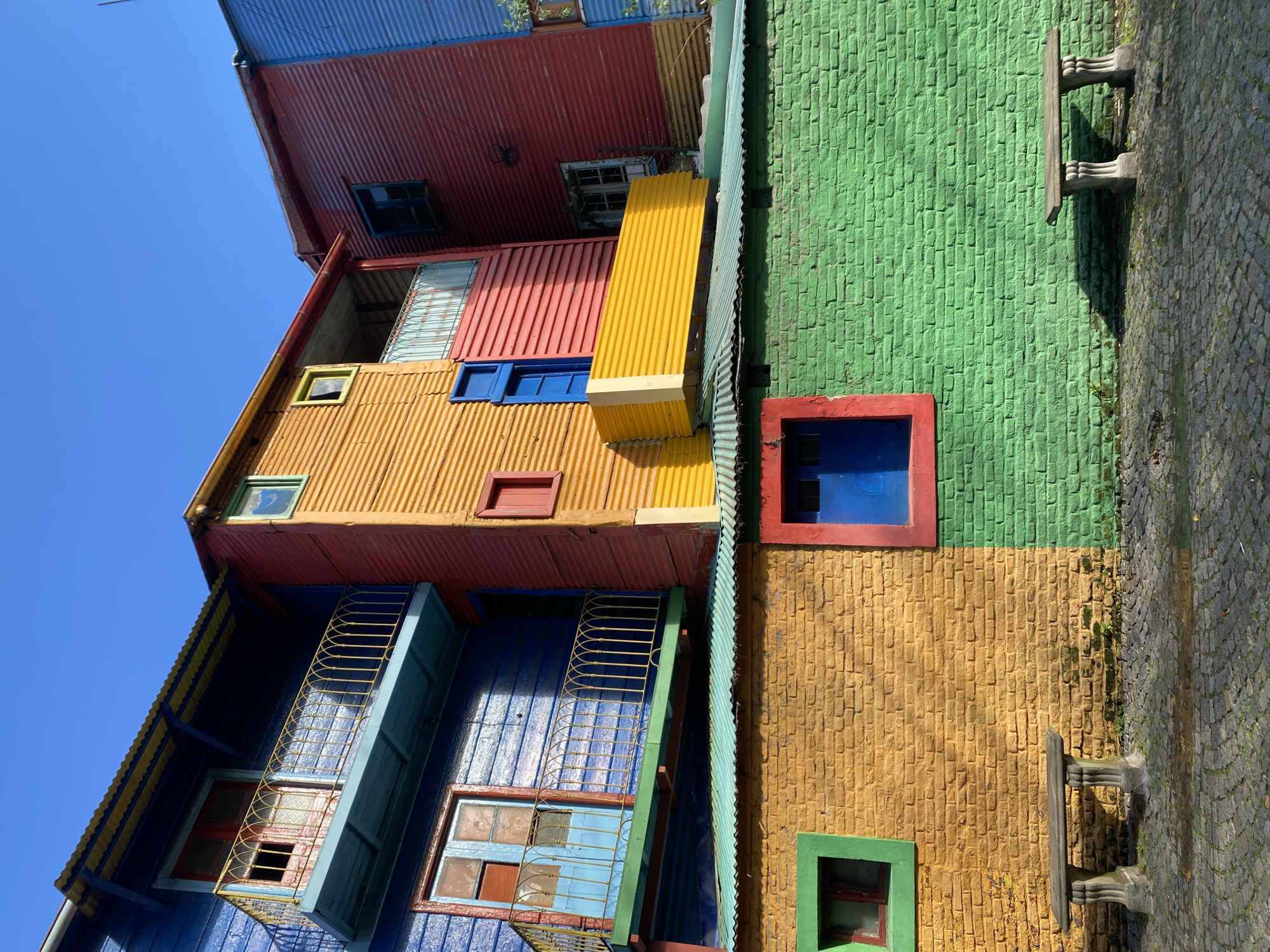This media file presents a vibrant and colorful street scene in Buenos Aires, Argentina, characteristic of the famous Caminito area in La Boca. The image is dominated by brightly painted buildings under a clear, bright blue sky, suggesting a sunny day.
The architecture is eclectic and lively, featuring a mix of corrugated metal siding and brickwork facades. On the right, a building displays a textured facade that is green on the upper section and orange below, accented with a red-framed blue window and a green-framed window. Adjoining this, a cobblestone street or walkway is visible, where two sturdy, rustic concrete benches or tables are placed, casting sharp shadows from the strong sunlight.
In the center, a prominent building is clad mainly in yellow corrugated metal, featuring several blue-framed windows, a small red-framed window, and a distinctive yellow horizontal protrusion, possibly an awning or part of a balcony structure. Sections of red corrugated metal are visible on its upper parts and on adjacent structures. Further to the left, another building exhibits a blue corrugated metal exterior with a yellow-railed balcony and multiple windows. The roofs of the various buildings are also made of corrugated metal, painted in shades of red and blue.
No people are present in the scene, which appears static, emphasizing the architectural and chromatic richness. There is no visible text. The overall impression is one of a cheerful and visually striking urban landscape.