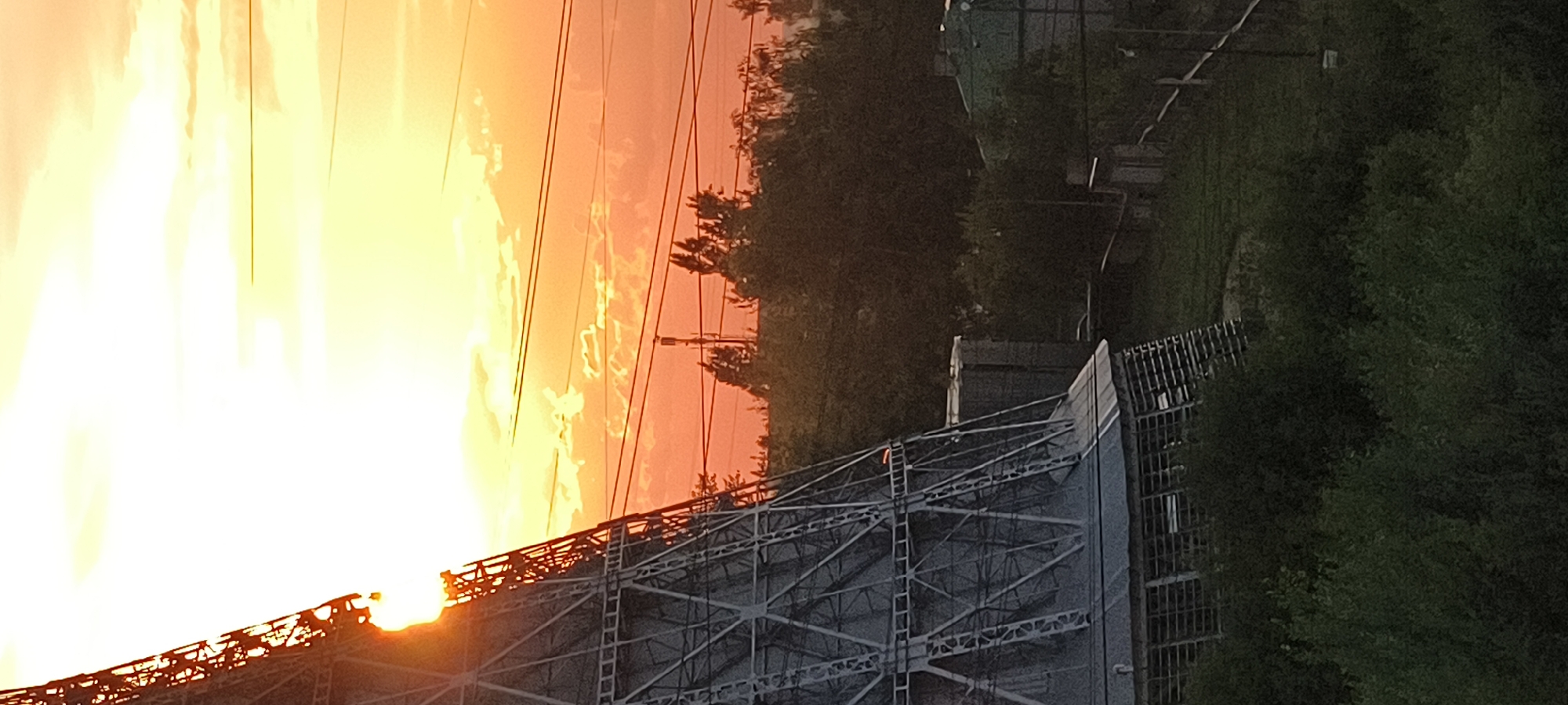 A wide shot captures a sunset over a landscape in an unknown city. The sun is a bright orange orb in the upper left corner, partially obscured by a large, grey, metallic structure that resembles a cooling tower or radio tower. The structure is composed of a lattice of beams and supports, with a sloping roof-like section at its base. The sky is filled with vibrant orange and yellow hues, with wispy clouds scattered across it. Several power lines stretch horizontally across the frame, contrasting with the warm tones of the sunset.
In the middle ground, a line of trees with dark green foliage creates a dense backdrop. The trees are varied in height and shape, with some taller specimens interspersed with smaller bushes. A few buildings are visible peeking through the trees, including a structure with a green roof and a darker, rectangular building adjacent to the large metallic structure. A wire fence runs along the edge of the property, bordering a grassy area. The foreground is dominated by lush green bushes and foliage, which obscures the lower portion of the scene. The overall impression is one of an industrial or utility area set against a dramatic natural backdrop of a sunset.