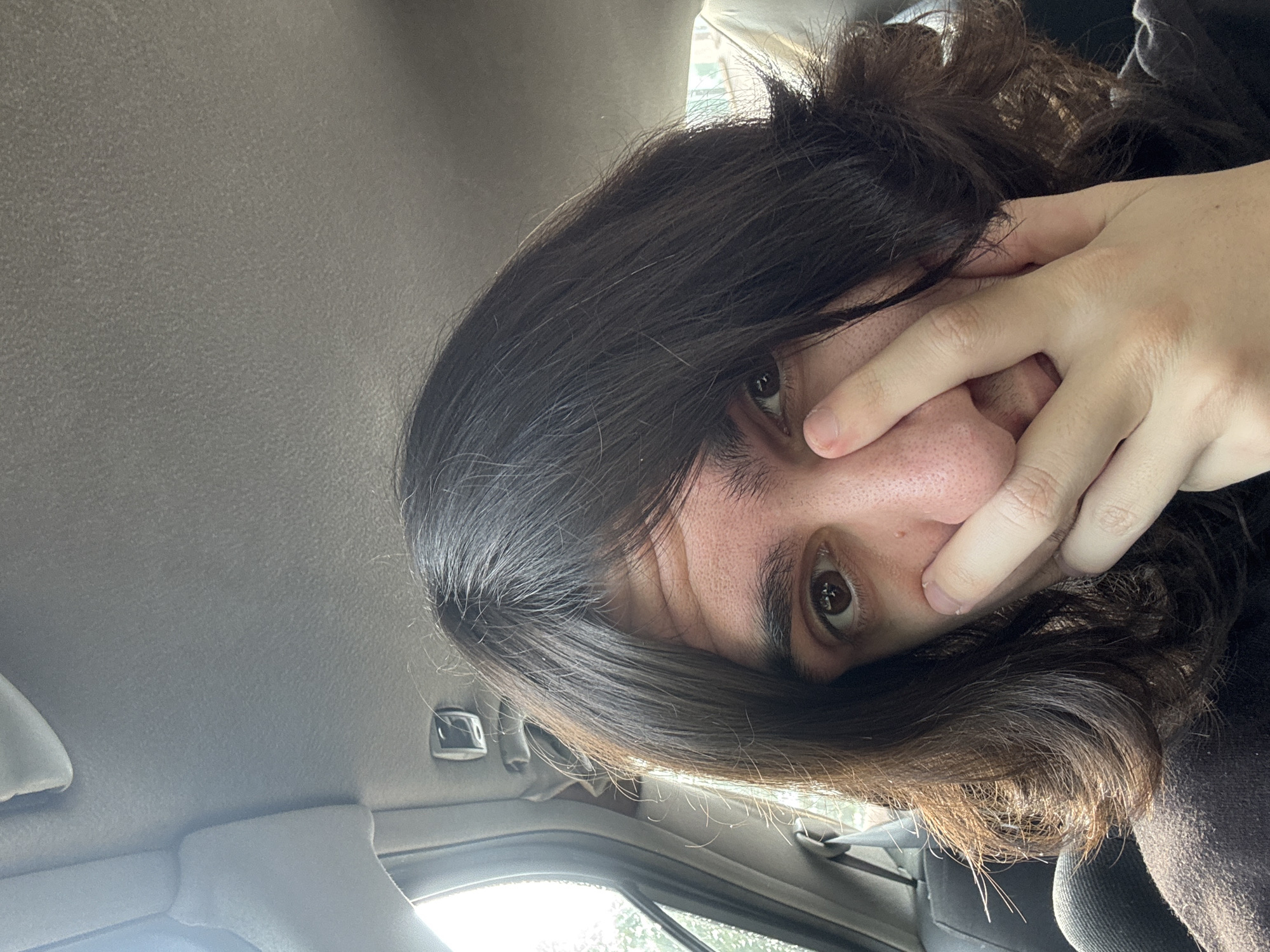 A rotated close-up portrait shows a person with long, dark brown hair partially obscuring their face with their hand, revealing one brown eye. The person is positioned inside a car, likely in Radnevo, Bulgaria, with their head towards the bottom-right of the frame, looking upwards. Their hand covers their nose and mouth, lending a sense of shyness or a casual, candid moment.
The car's interior features a light grey textured headliner, visible across the top and left of the frame. Bright daylight streams in from a side window, indicating it is daytime. Parts of the car's window frame, a headrest, and interior fittings are also visible. The person is wearing a dark, possibly black or dark grey, top. No specific activity other than posing for the photograph is depicted. No text is visible in the image.