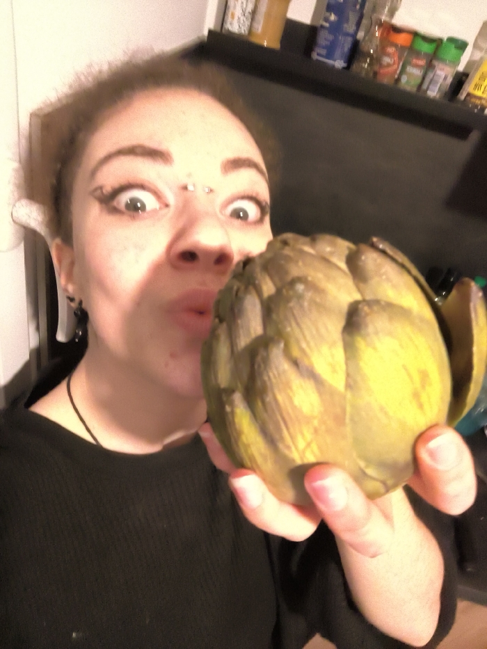 A woman holds a large artichoke close to her face, pulling a funny expression with wide eyes and pursed lips. She wears a black shirt, a necklace, and has visible eyebrow piercings. Her hair is light brown and curly. In the blurry background, a shelf displays various spices and jars. The lighting appears to be artificial, with a slight yellow tint.