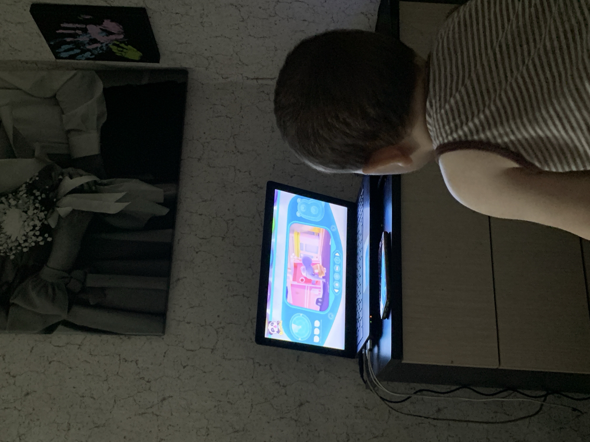 A child is looking at a wall-mounted digital device showing a cartoon. To the left of the device are two pictures on the wall. One appears to be a handprint in paint and the other appears to be a black-and-white wedding photograph. The location is not discernible from the given image.