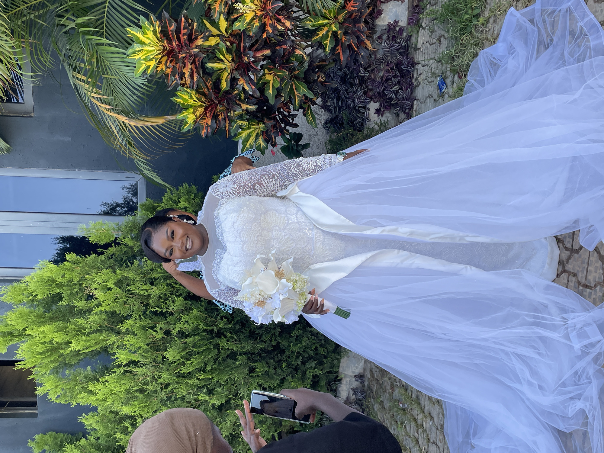 A woman in a white wedding dress is photographed in Jos, Nigeria. She holds a bouquet of white flowers. Another person is taking a picture of her with a cell phone. They are outside in a garden with various plants.
