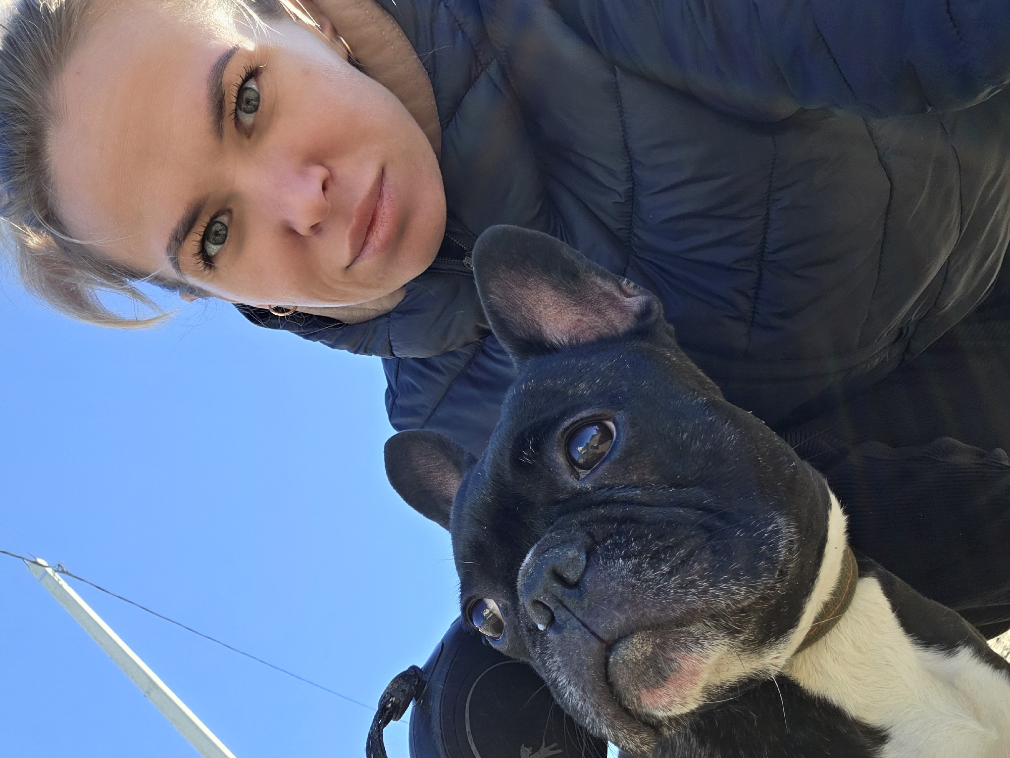 The image shows a woman and a black and white French Bulldog. The woman is looking at the camera with a slight smile. She has light hair and green eyes. She is wearing a black puffer jacket. The dog is looking to the side. It is wearing a brown collar. The background is a clear blue sky. A white mast and line are visible in the lower left corner of the image. Based on these features, the location is possibly in Chaikovsky, Russia.