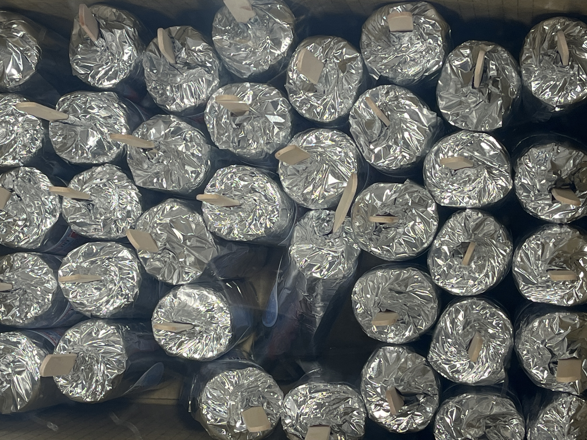 A top-down, close-up shot presents a dense arrangement of numerous cylindrical objects, tightly packed into what appears to be a container or box. Each object is individually wrapped in highly reflective, crinkled silver foil, likely aluminum. A small, flat, light-colored wooden stick or tab protrudes vertically from the center of the foil-wrapped top of many of these items.
The objects are arranged in close rows, filling the entire frame. The crinkled texture of the foil is prominent, reflecting light unevenly and creating bright highlights and deep shadows across the surface. The areas visible between the wrapped items and at the edges of the container are dark, suggesting a dark background or the interior of a box. The combination of foil wrapping and wooden sticks strongly suggests these items are frozen treats like ice cream pops or similar confections.
There is no discernible activity taking place, only the static display or storage of these products. No people are visible. While most surfaces are silver and light wood, some faint, blurry text or markings, appearing as dark or reddish smudges, can be seen beneath the foil on a few items, though it is illegible. No specific location, time of day, or weather can be inferred from the visual information alone.