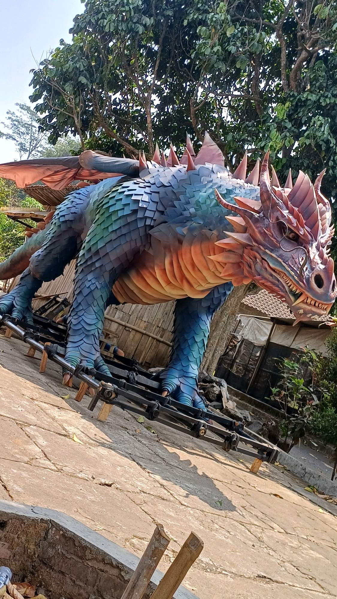 In Karangwuni, Indonesia, a large, vibrantly colored dragon sculpture is positioned outdoors. The sculpture features blue and green scales on its body, with orange and red accents on its underside and head. It has prominent horns, sharp teeth, and visible claws. The dragon is elevated on a black metal and wood support structure, which appears to be some form of scaffolding or mechanical rig. Behind the sculpture, there are trees with dense green foliage and a partially visible building with a tiled roof. The ground is a textured concrete surface, with cracks and some debris, including fallen leaves. The lighting suggests it is daytime with bright sunlight casting shadows.