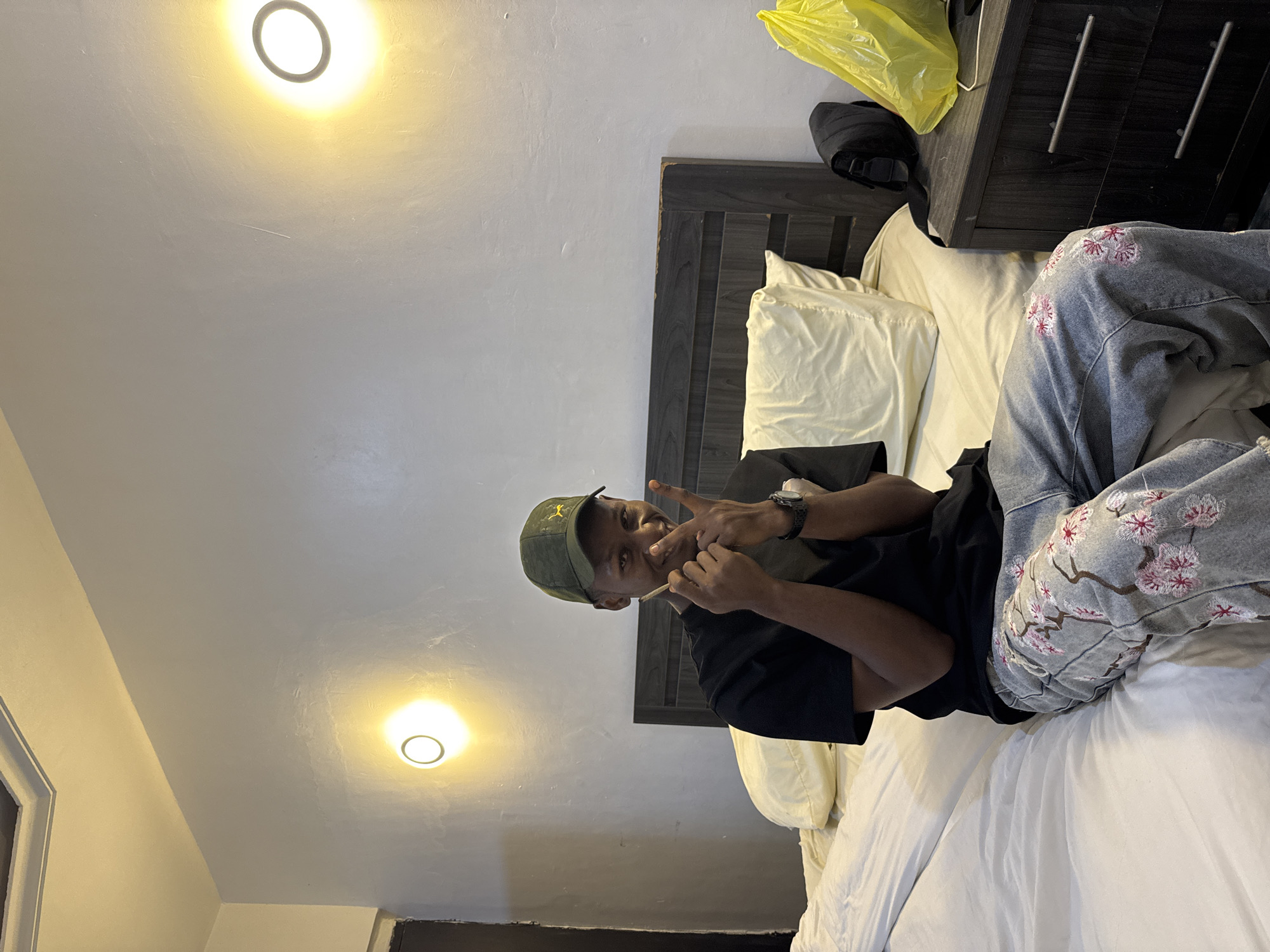 A person is lying on a bed in a room. The person is wearing a black shirt, jeans with floral embroidery, and a green cap. The person is holding a rolled cigarette in their hand. The room has white walls, a dark wooden headboard, and a nightstand with a yellow plastic bag on it. There are two light fixtures on the ceiling. The location is not specified in the image, but I am instructed to say it is in Awka, Nigeria.