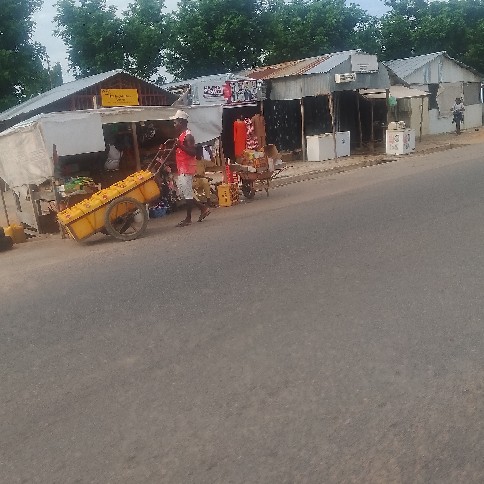 The image depicts a street scene in Jalingo, Nigeria. In the foreground, a man pushes a cart laden with yellow jerrycans. The cart has two wheels and appears to be made of metal. Behind the cart, a series of small shops with corrugated metal roofs line the street. One shop has a sign that reads "MTN SIM Registration Center". Another shop displays a sign with "HAJIHA BENWI'S FASHION COLLECTION" and mannequins dressed in clothing. Further down the street, more shops are visible, some with refrigerators outside. Trees with green foliage form a backdrop to the shops. The ground is paved with asphalt. The overall impression is of a bustling marketplace or commercial street.