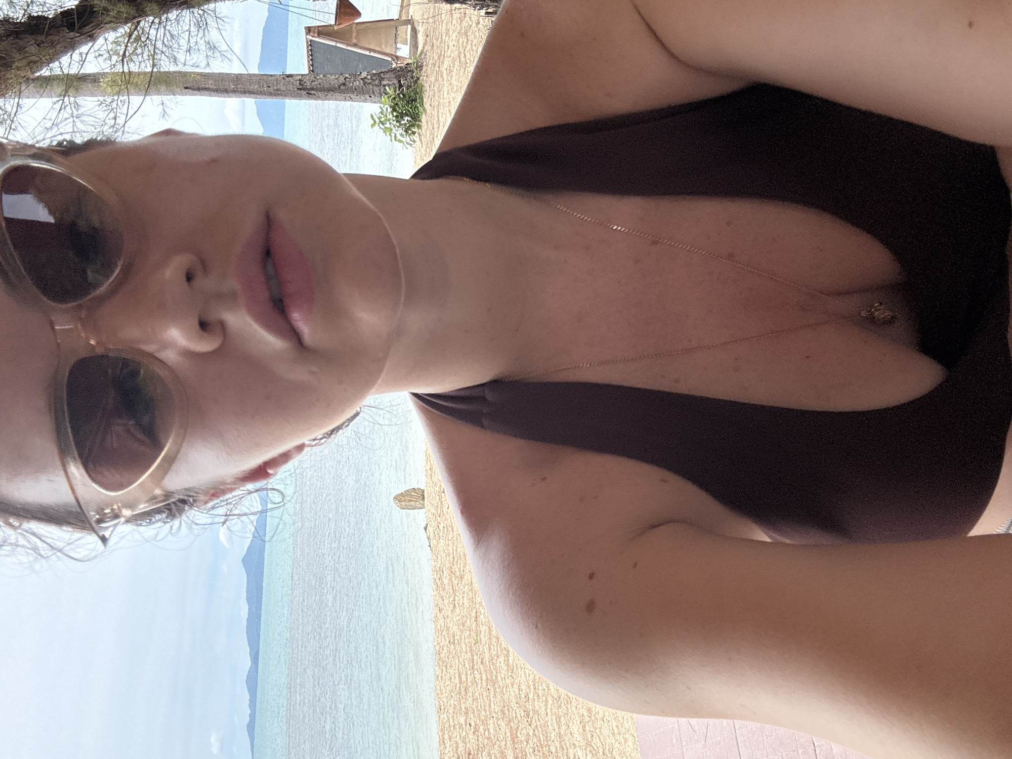 A woman is taking a selfie on a beach in Baan Tai, Thailand. She is wearing sunglasses and a dark brown bikini top. The ocean and distant islands are visible in the background, along with palm trees and a small structure. The beach appears to be made of light-colored sand.
