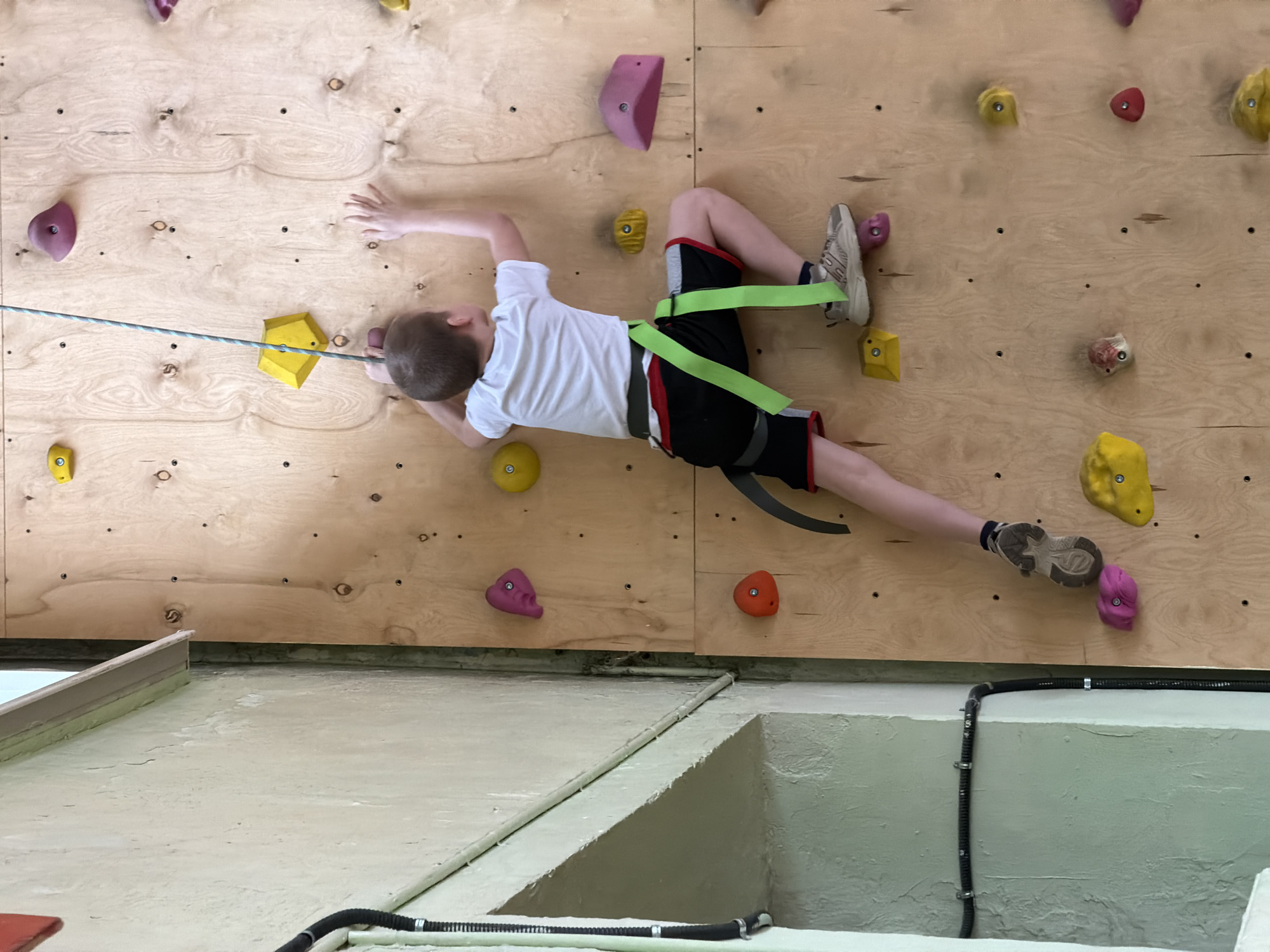 A child wearing a climbing harness and sneakers is climbing a climbing wall inside a building. The wall is made of plywood and has various colored climbing holds attached to it. A rope is attached to the child's harness. The building is in Izhevsk, Russia. A portion of a wall is visible below the climbing wall. A cable conduit runs along this wall.