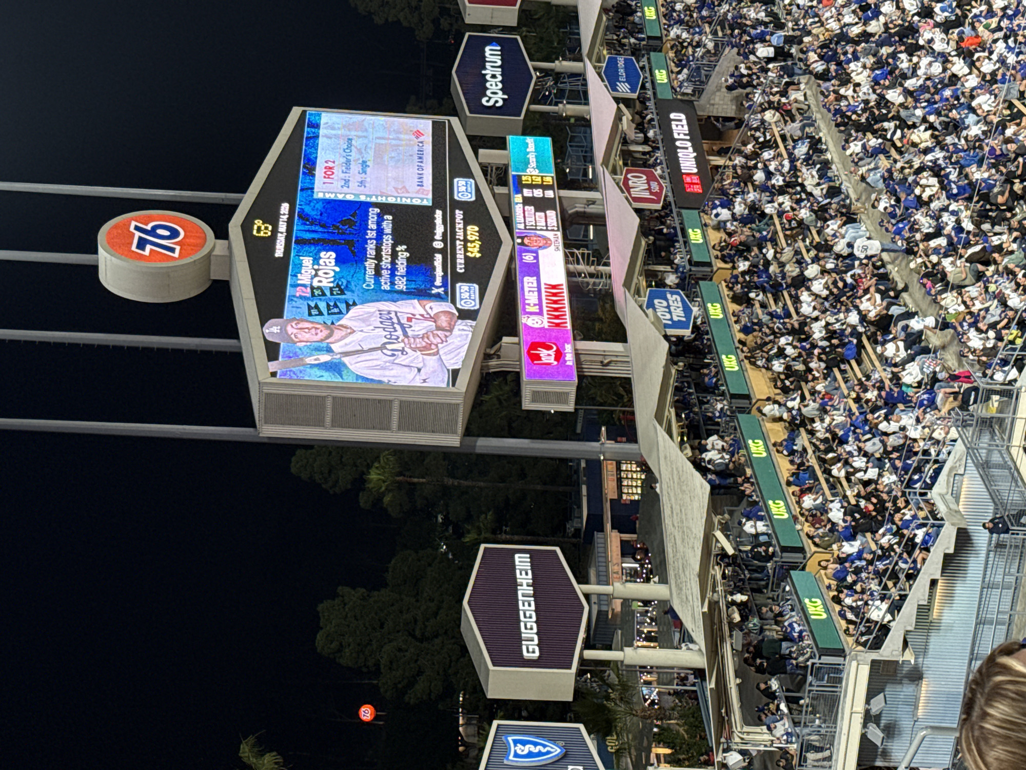 This image shows a night scene at Dodger Stadium in Los Angeles. A large crowd of people, many wearing blue and white, fills the stadium seats. Advertisements are visible on the stadium signage including Spectrum, Uniqlo Field, Toyo Tires, and Guggenheim. The scoreboard features information on Miguel Rojas and a 50/50 jackpot, along with the temperature of 63 degrees and the date of Thursday, July 14, 2026. A "76" gas station sign is also visible.