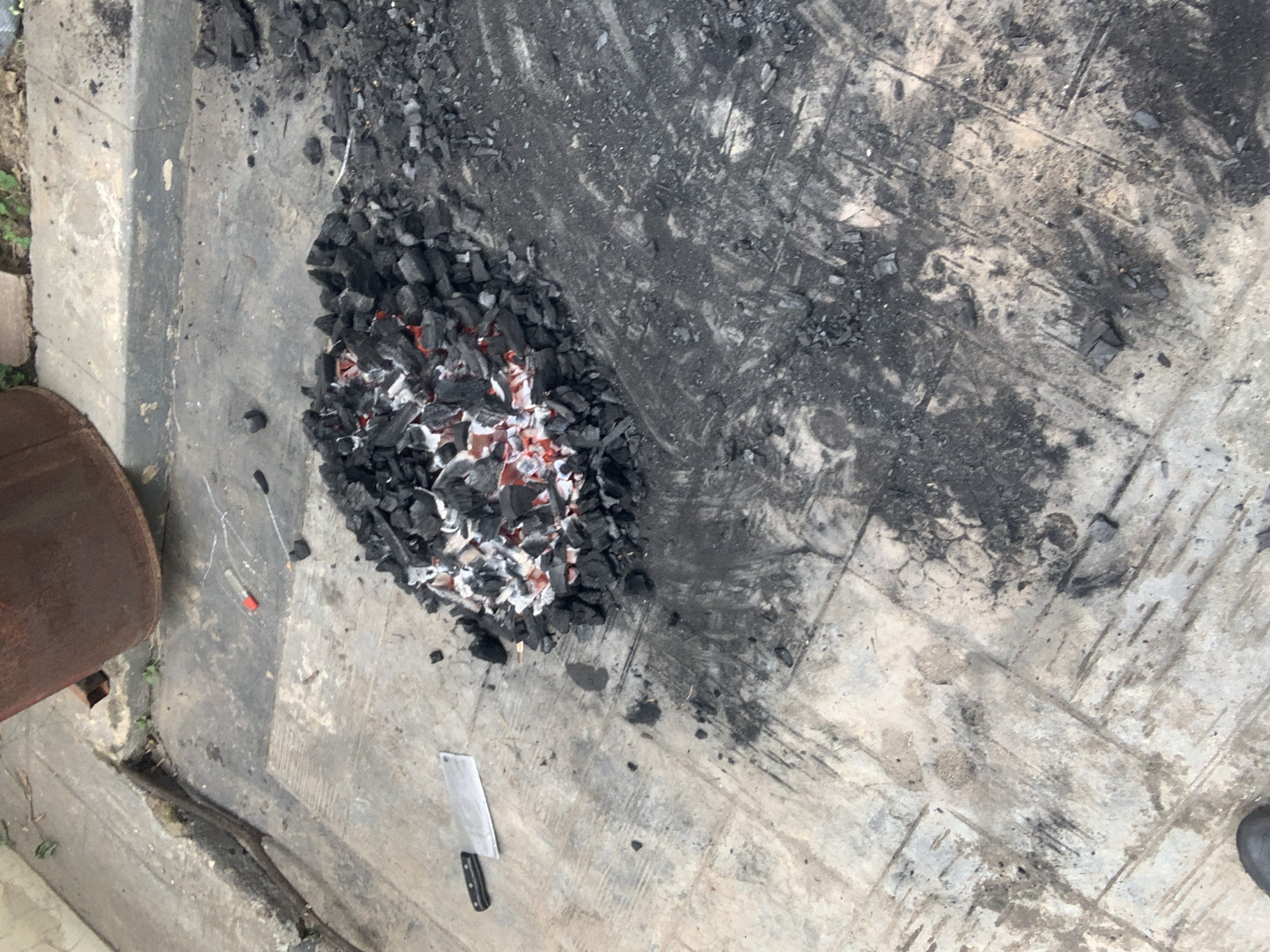 The image depicts a pile of glowing charcoal on a concrete surface, possibly in an outdoor setting within Abuja, Nigeria. The charcoal is centrally located, with some scattered around it. The concrete surface appears weathered and stained, with visible cracks and dark patches indicating past fires or spills. To the left of the charcoal pile, a cleaver is placed on the concrete, its metallic blade glinting. A lighter with a red cap is also visible nearby. In the background, a large, rusty barrel is partially visible, suggesting it might have been used in conjunction with the fire. The overall environment appears to be an industrial or a utilitarian outdoor space. There are no people visible in the image.
