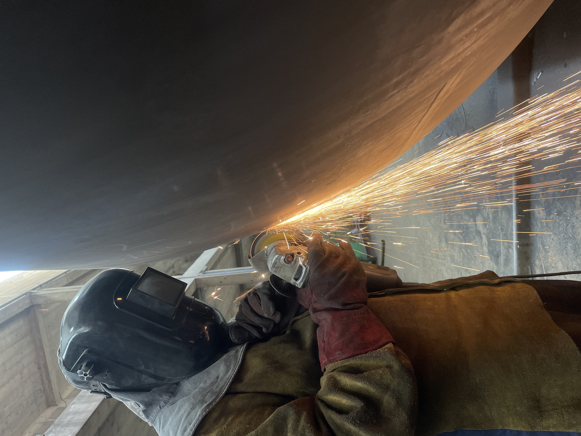 A person is welding a large, dark metal structure in what appears to be an industrial setting. The welder is wearing a protective helmet, gloves, and a heavy apron-like garment. A shower of sparks is emanating from the welding point. The surrounding environment has exposed concrete beams, suggesting a warehouse or similar construction site. The location is Čeluga, Montenegro.