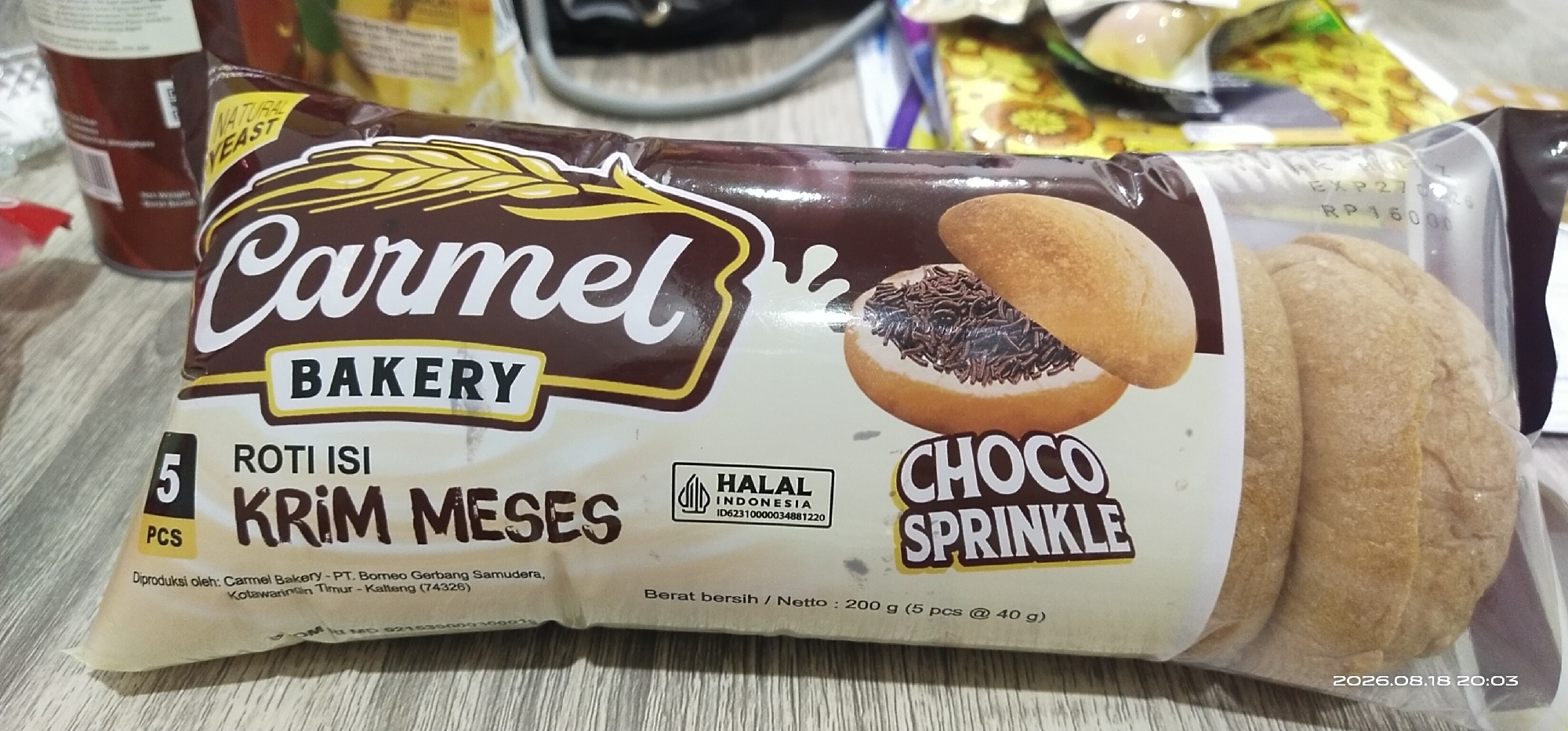 The image shows a package of "Carmel Bakery Roti Isi Krim Mesis Choco Sprinkle" bread rolls. The packaging is primarily brown and white, featuring a stylized image of wheat stalks, the Carmel Bakery logo, and an illustration of a bread roll filled with cream and chocolate sprinkles. Text on the packaging indicates "NATURAL YEAST," "5 PCS," "ROTI ISI KRIM MESES," "HALAL INDONESIA," and "CHOCO SPRINKLE." It also states the net weight is "200 g (5 pcs @ 40 g)" and that it is "Diproduksi oleh: Carmel Bakery - PT. Borneo Gerbang Samudera, Kotawaringin Timur - Kalteng (74326)." The packaging also displays an expiration date of "EXP 27/02/2026" and a price of "RP 16000". The bread rolls themselves appear golden brown and are visible through the clear plastic packaging. The background consists of a wooden surface, likely a table. The image is a close-up of the product, with no indication of a specific location within the city of Matabu, Indonesia, beyond the manufacturing address.
