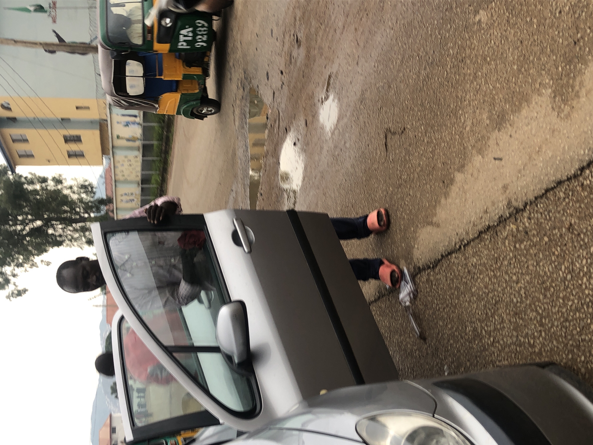 In Jos, Nigeria, a man in a collared shirt and dark pants, wearing red slip-on sandals, stands by an open car door. Another person, wearing a red garment, is visible in the passenger seat through the open door. A three-wheeled auto-rickshaw with the license plate "PTA 9289" drives past on a wet, asphalt road, reflecting the sky. In the background, a yellow and blue building with arched windows is visible, and a mural depicting figures can be seen on a wall. Trees and a hilly landscape are present in the distance. The scene appears to be outdoors, with puddles on the road suggesting recent rain.