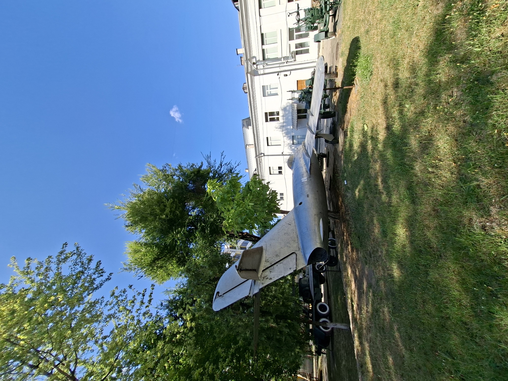 An outdoor scene in Chișinău, Moldova, features a weathered, silver-colored jet airplane displayed on a grassy lawn in front of a white, multi-story building with numerous windows and a flat roof. The aircraft, positioned at an angle, exhibits signs of age and exposure to the elements. Its tail fin is prominently visible, pointing upwards. Behind the jet, a dark-colored truck with dual rear wheels is partially visible. To the right of the jet, a green artillery piece on a wheeled carriage is also on display. Lush green trees with dense foliage flank the left side of the scene, casting dappled shadows on the grass. The sky above is a clear, bright blue with a few wispy clouds. The overall impression is that of an outdoor museum or exhibition space.