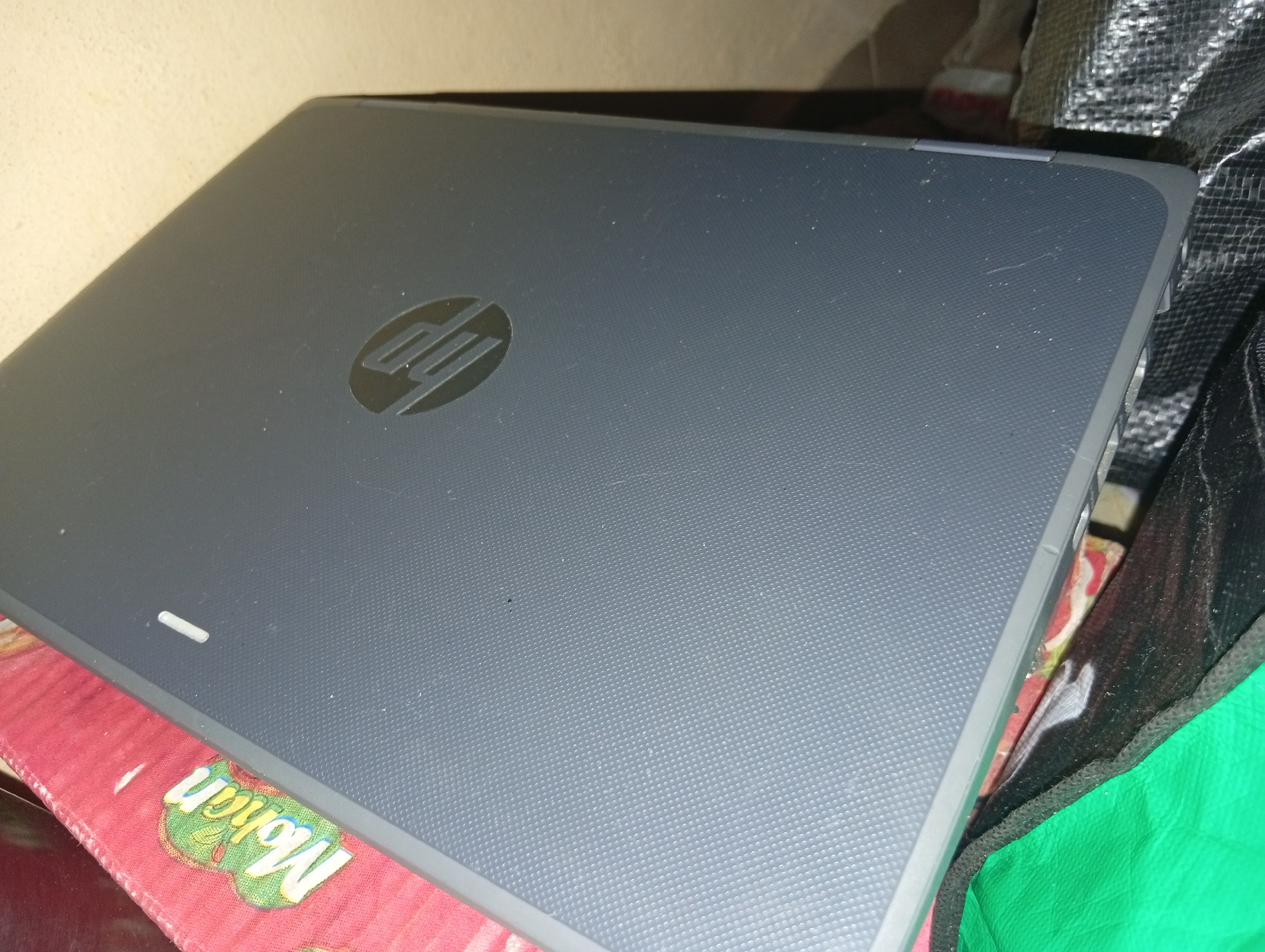 The image shows the closed lid of a dark gray laptop with the HP logo imprinted in the center. The laptop rests on a red, patterned cloth with green and yellow designs, possibly a mat or a tablecloth. To the right of the laptop, a black mesh bag with a green lining is visible, and behind the laptop, a portion of a wall or partition can be seen. There is no indication of Ukukwa, Nigeria in the image itself.
