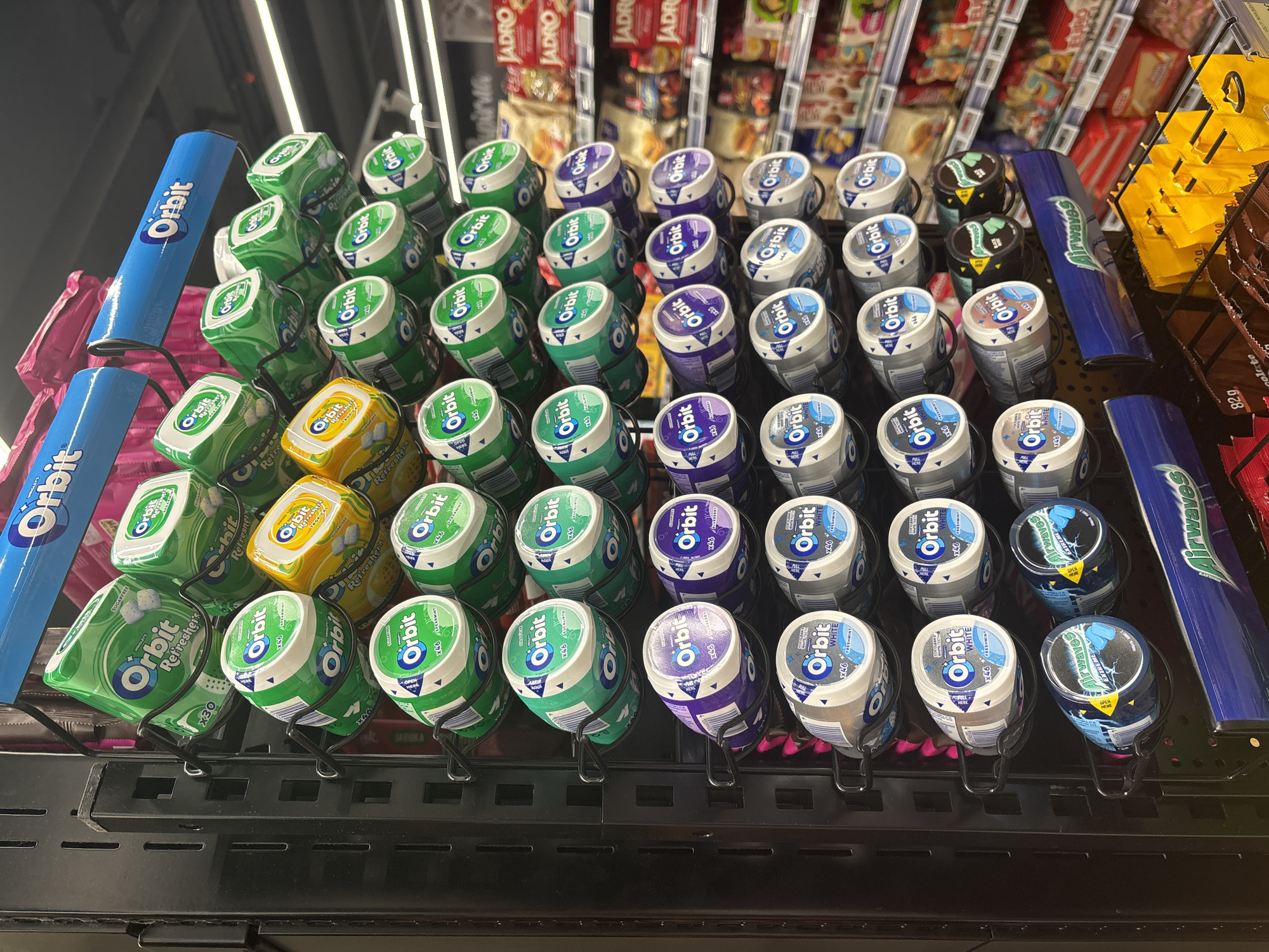 The image presents a close-up view of a candy rack in what appears to be a retail environment, possibly a grocery store or convenience store. The focus is on a selection of Orbit brand chewing gum in various flavors and packaging. The gum is arranged in rows on a metal rack, with each container individually held in place. The color palette includes shades of green, blue, yellow, purple, and white, corresponding to different gum flavors. Brands alongside Orbit chewing gum include Airwaves gum, and JADRO snack. The rack is situated under fluorescent lighting. It is impossible to discern the location of this image in Budva, Montenegro, or if the image was taken there, without additional information. There are no people visible in the image.