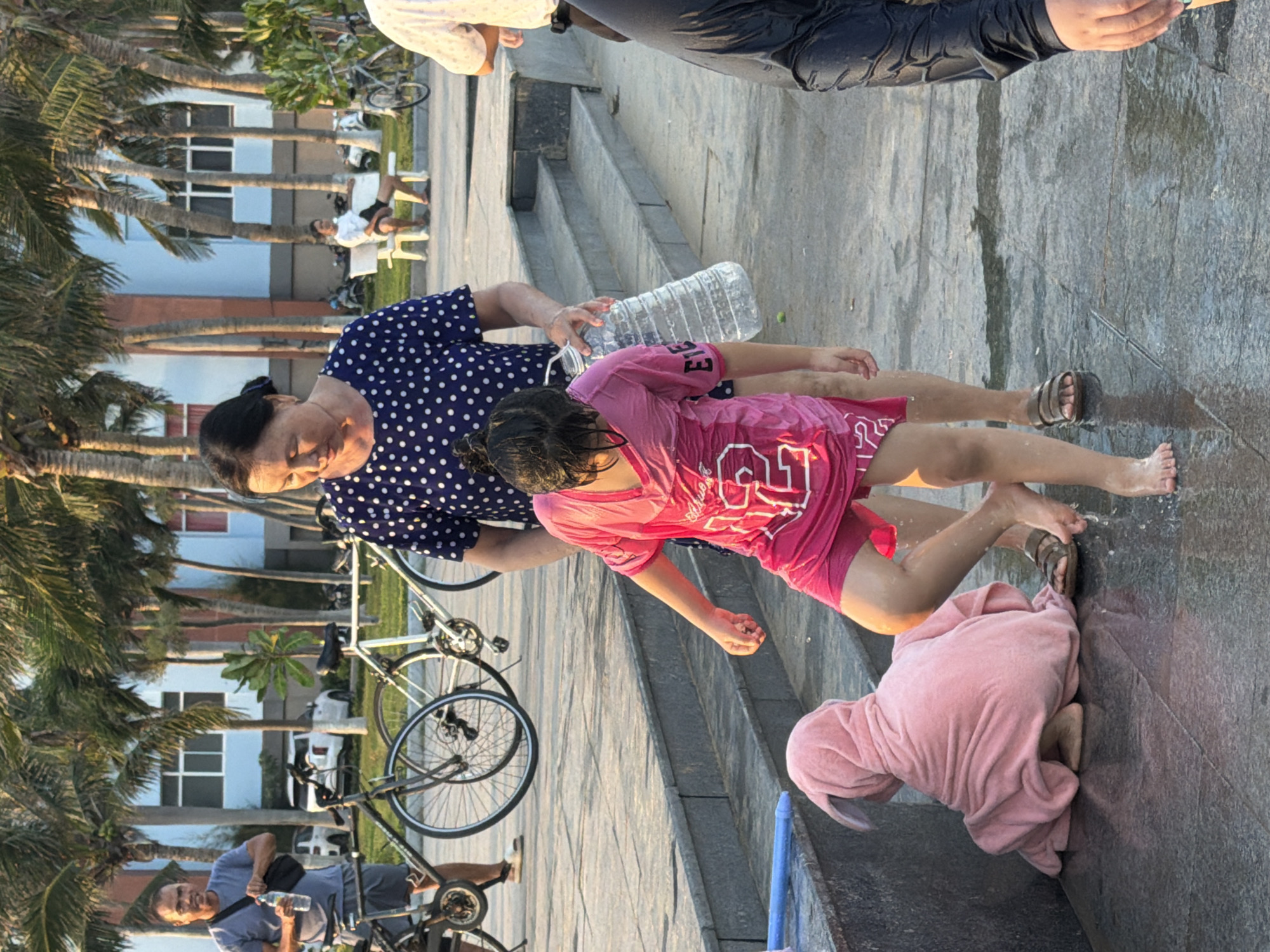 In the city of South Quy Nhon Ward, Vietnam, a woman in a blue and white polka dot dress stands holding a large plastic bottle. Beside her, a young girl in a wet pink t-shirt with white lettering and pink shorts splashes water. To the left, a child wrapped in a pink towel with a bunny-like hood sits on the wet ground. In the background, palm trees line a pathway with bicycles and buildings. A man in a grey t-shirt and shorts stands by a bicycle on the left, and another person in a white shirt is visible on a bench in the distance on the right. The scene appears to be outdoors on a paved area with some wet surfaces, suggesting a recreational or public space.