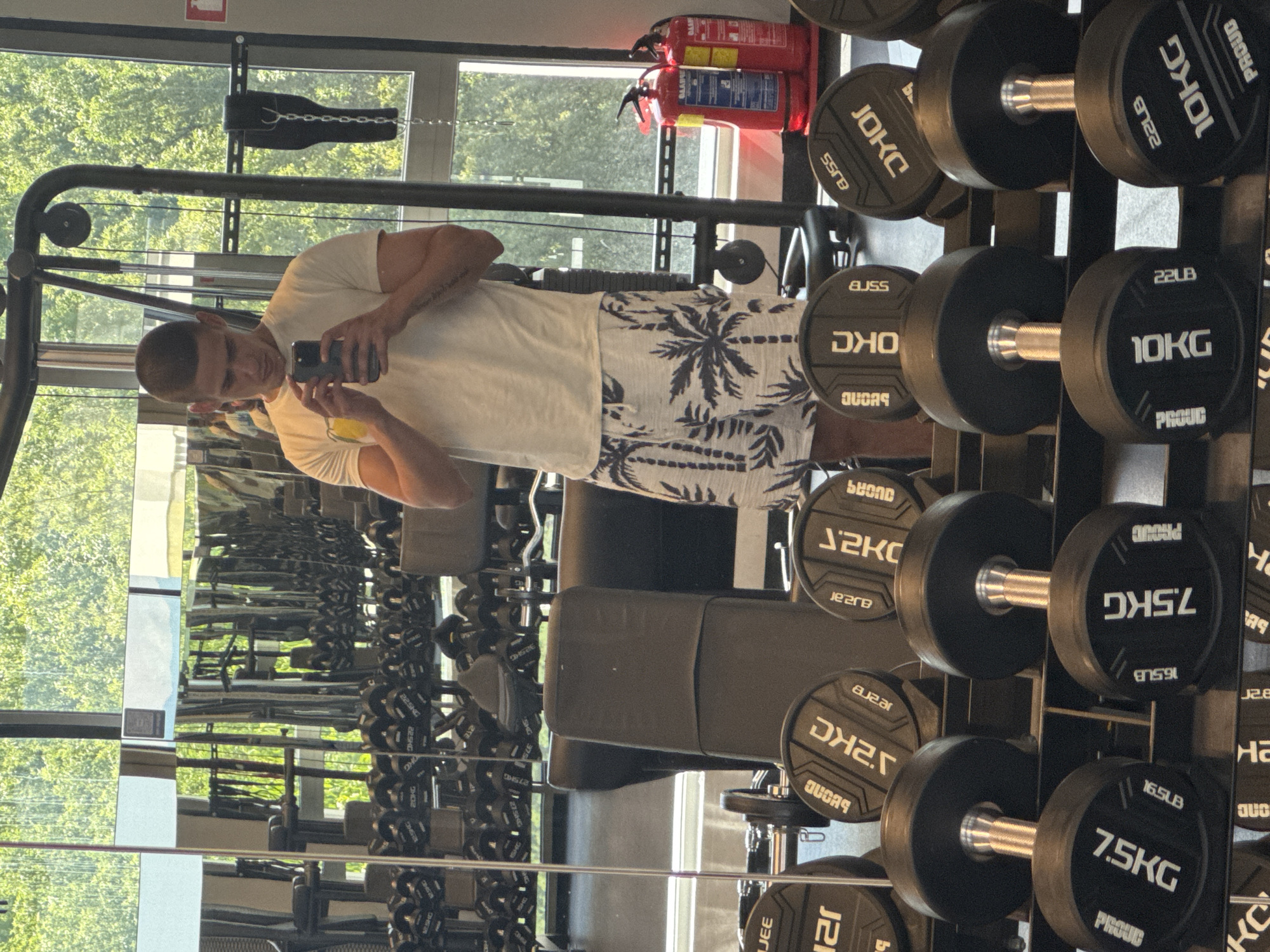 A young man, with short dark hair and a tattoo on his right arm, stands in a gym, taking a selfie in a mirror. He is wearing a light-colored t-shirt and white shorts with a palm tree pattern. In front of him, a rack of dumbbells is visible. The gym features large windows, allowing natural light to illuminate the space and offering a view of trees and greenery outside, suggesting an urban location in Sofia, Bulgaria. Two red fire extinguishers are mounted on a wall to the right of the frame. The overall setting is a well-equipped fitness facility.