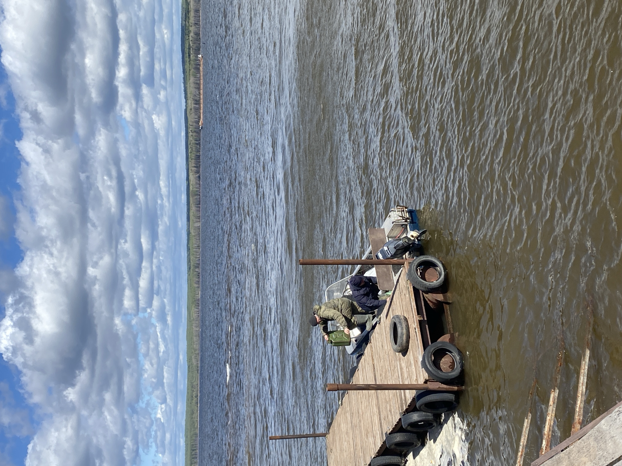 A medium-angle shot depicts a wooden pier extending into a body of water, where a small boat is docked. Two people are on the boat; one is bent over and appears to be handling a green container. The second person is seated in the boat's rear. The pier is lined with tires as bumpers. Metal posts support the pier. The water surface is textured with gentle ripples. In the background, a strip of land is visible with trees, and above, the sky is filled with white, puffy clouds against a blue backdrop. The image is taken outdoors. The city of Chaikovsky, Russia is not evident in the scene.