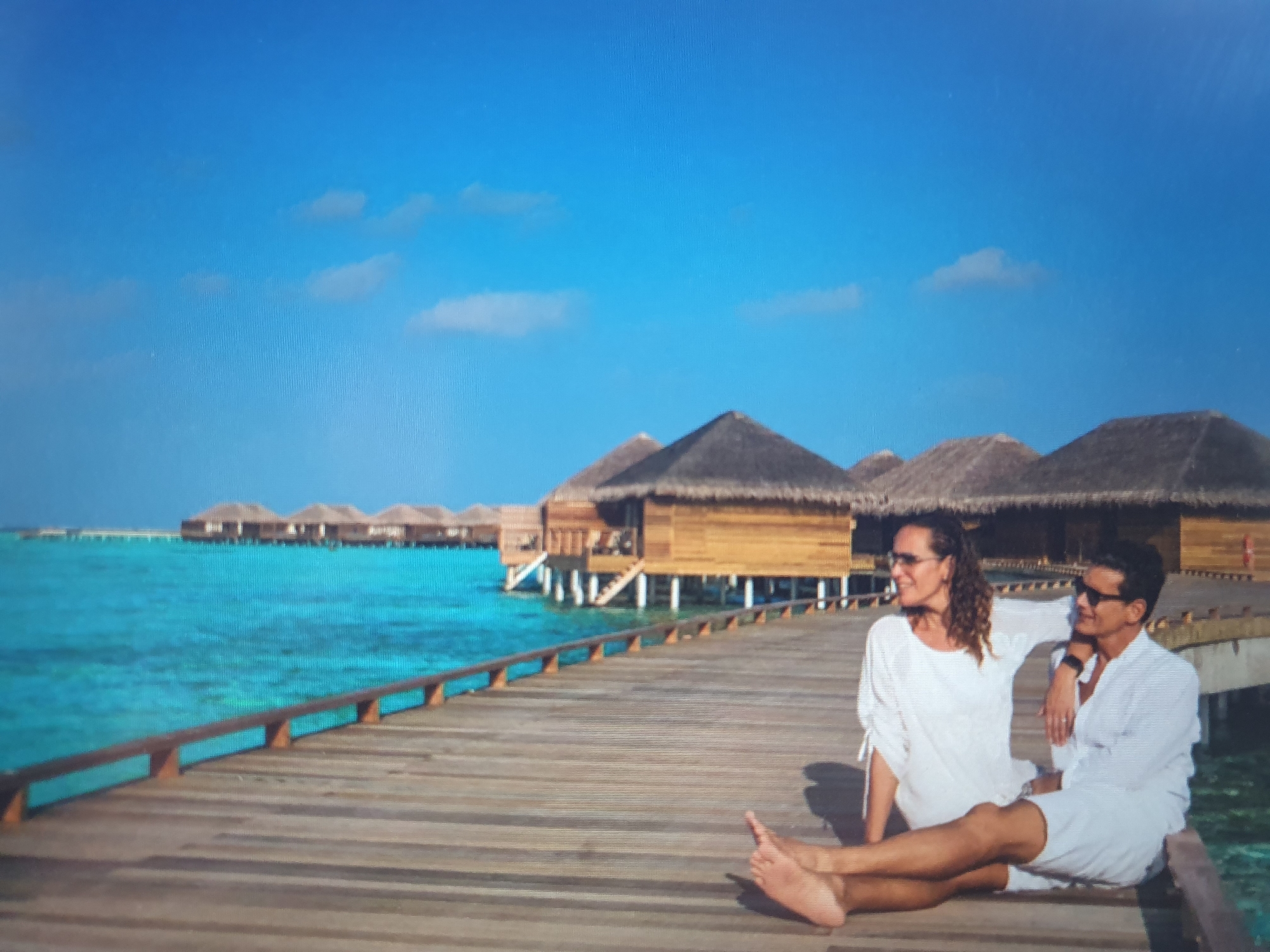 A couple is seated on a wooden pier overlooking a turquoise body of water. They are both wearing white clothing and sunglasses. The woman has curly brown hair and is leaning her head on the man's shoulder. The man is looking away from the camera. In the background, there are several overwater bungalows with thatched roofs. The sky is bright blue with a few scattered clouds. This image is not located in Tashkent, Uzbekistan.
