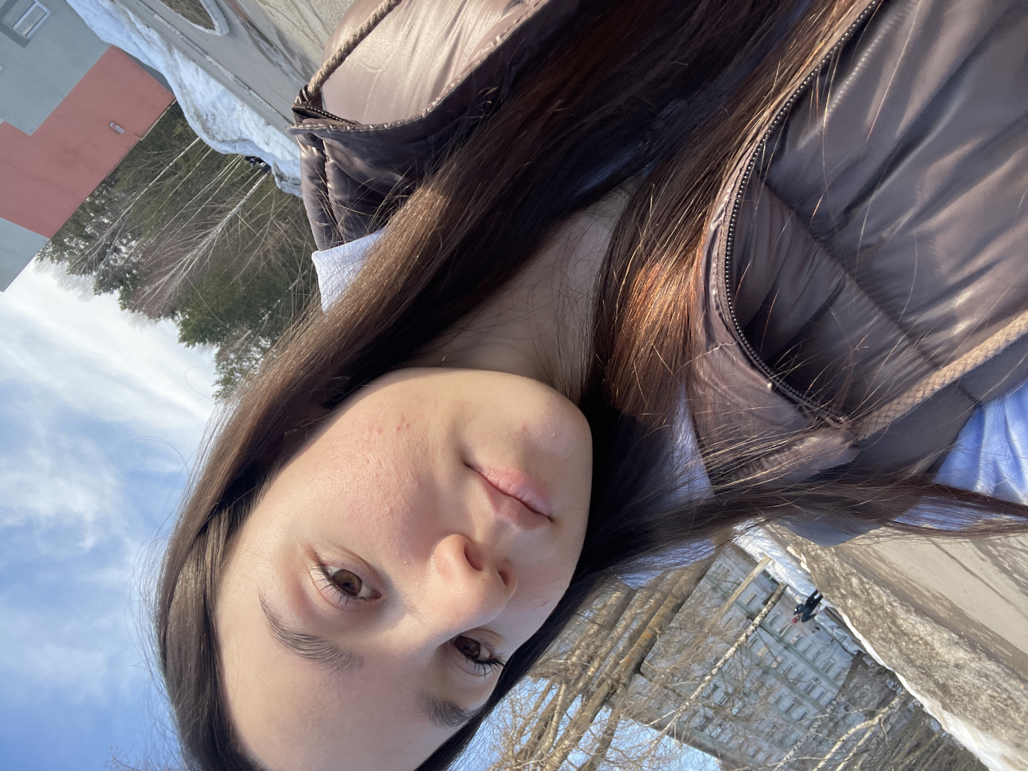 A young woman is taking a selfie in an outdoor environment. She has long, dark brown hair and is wearing a brown puffer jacket. The background shows a snowy street, a building with a red facade, and trees with bare branches. The location is Chaikovsky, Russia.