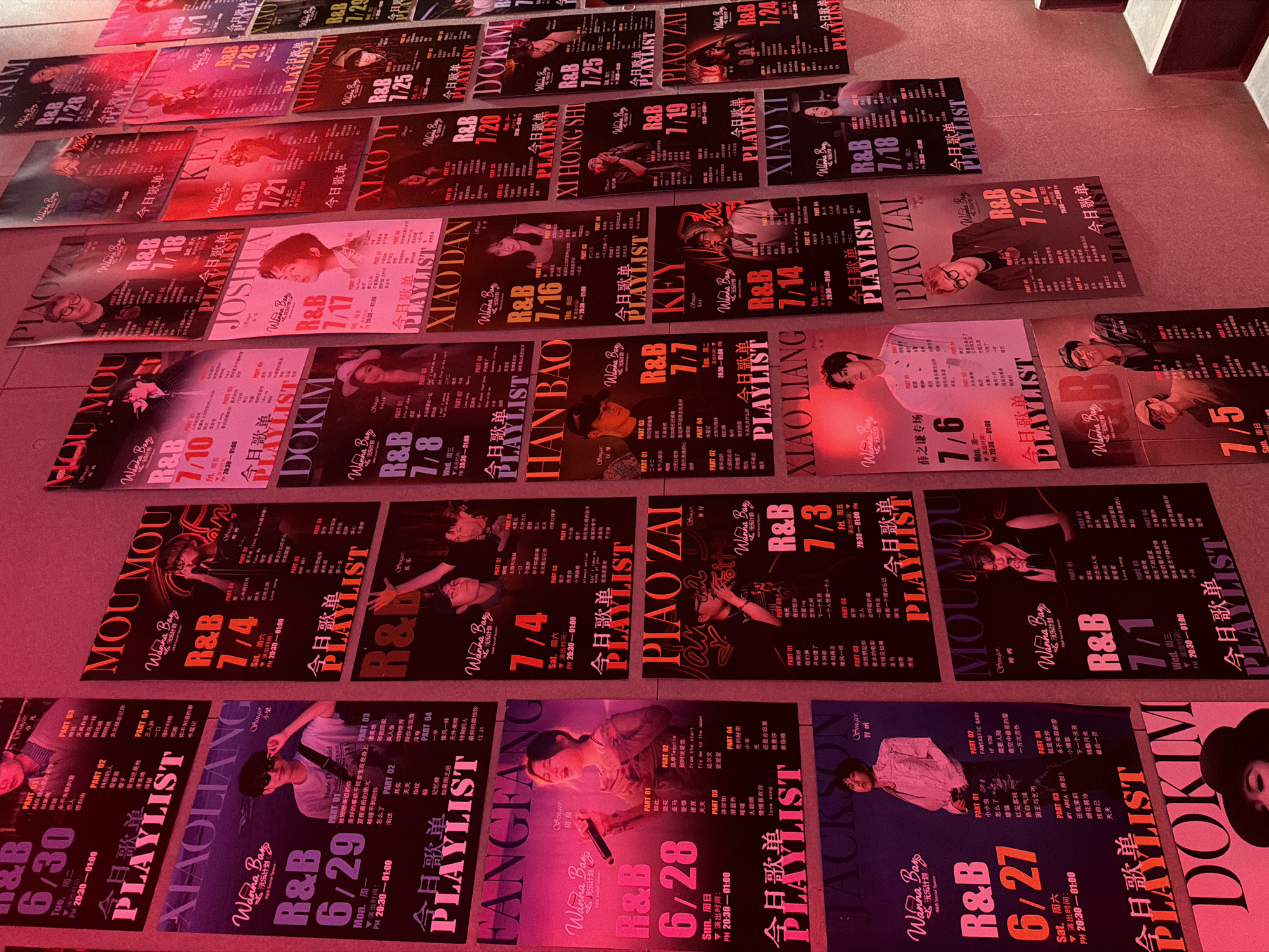 The image shows a wall covered with numerous posters, primarily for R&B music events. The posters feature images of various singers and groups, along with event details such as dates, times, and song titles. Many posters have the text "今日歌单 PLAYLIST" (Today's Song List PLAYLIST) prominently displayed. The posters are predominantly in shades of red and pink, suggesting a consistent theme or style. Some posters also include Chinese characters, indicating the location of these events. The overall impression is of a vibrant music venue or promotional display in a Chinese-speaking region. The lighting in the scene is warm and atmospheric, contributing to the energetic feel. The posters are arranged in a dense, overlapping manner on the wall, creating a visually rich and dynamic presentation. The city of Haizhu District, China is a known hub for entertainment and cultural events.