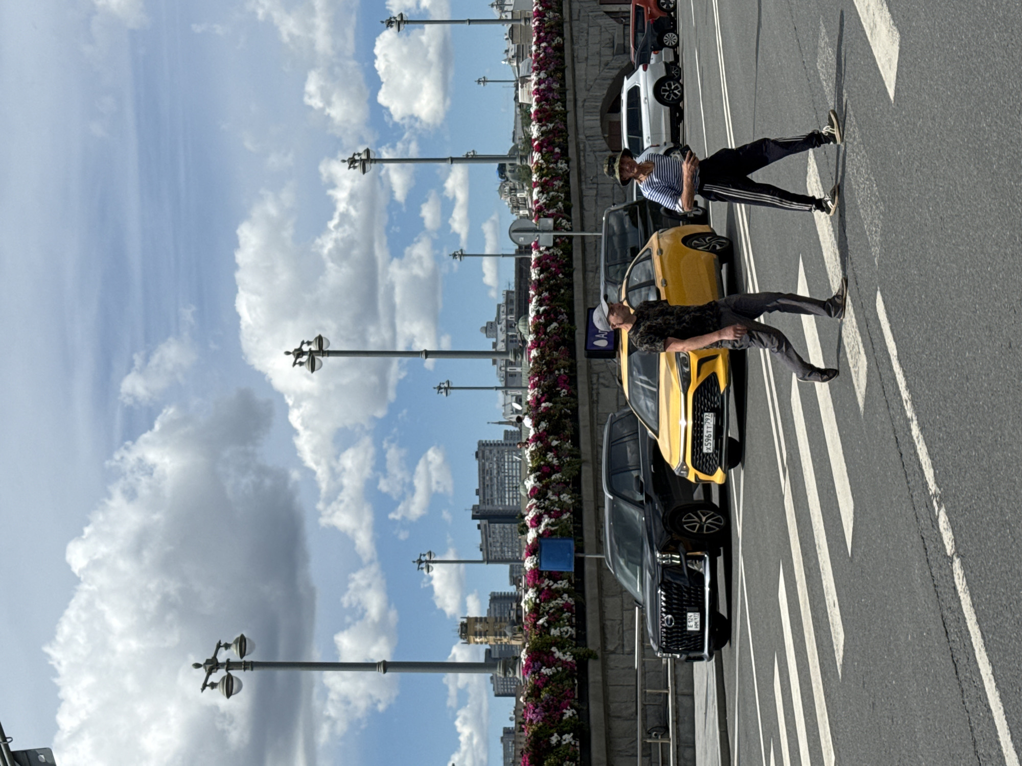 A wide shot captures a street scene in Moscow, Russia, on a partly cloudy day. In the foreground, a pedestrian crossing is marked by white stripes on asphalt. Two men are walking across the street on the pedestrian crossing. The man on the left is wearing a camouflage t-shirt, dark pants, and a grey baseball cap. The man on the right is wearing a striped shirt, dark track pants with white stripes, and a hat. Behind the men, a yellow car and a black SUV are stopped, presumably at a red light, with other vehicles further in the background. A decorative bridge or overpass with abundant pink and white flowers spans the background. Several street lamps and buildings are visible in the distance, under a sky with scattered clouds.