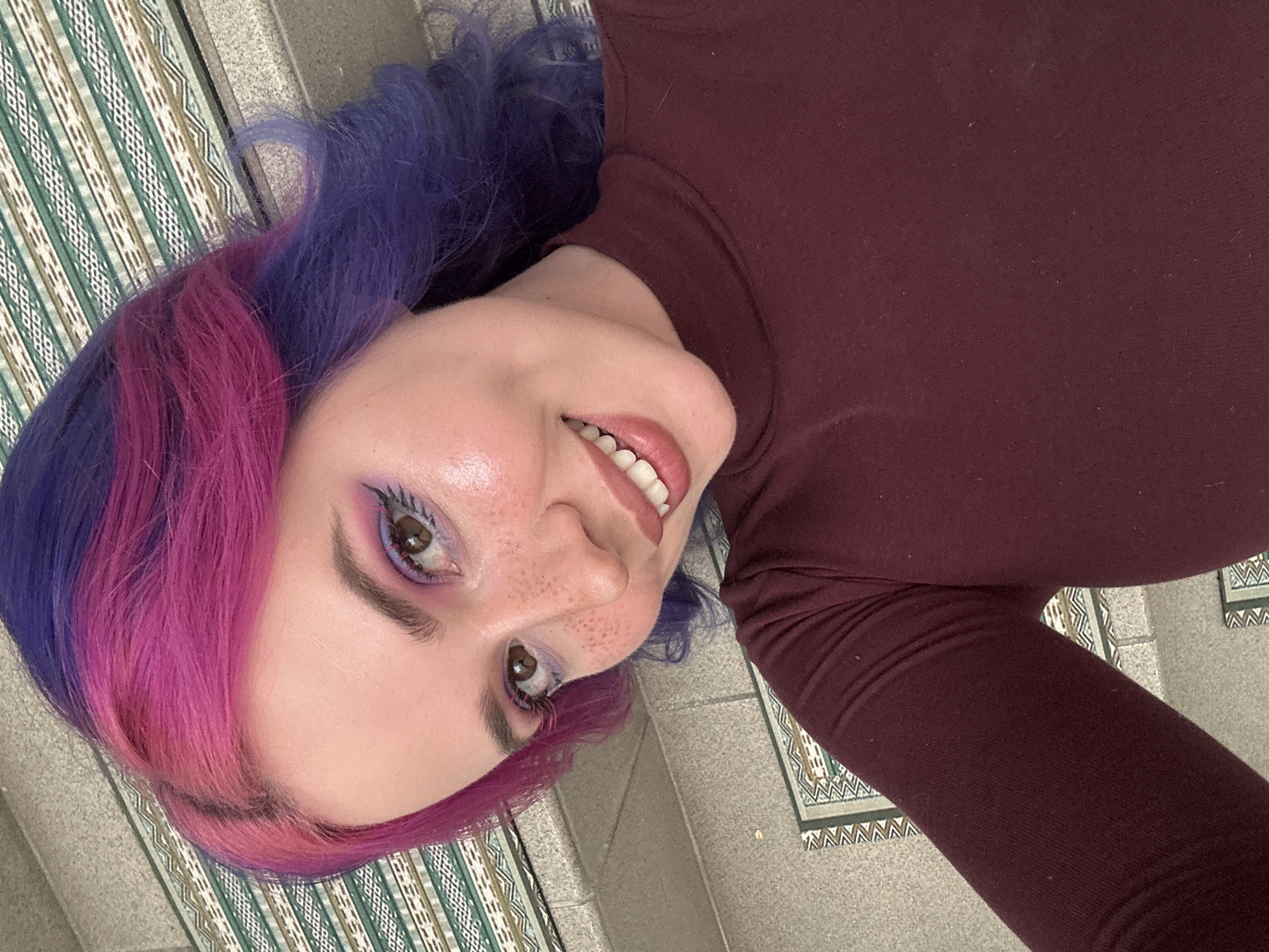 A close-up portrait shows a young woman lying down, looking up at the camera with a cheerful, open-mouthed smile. Her hair is strikingly colored with vibrant sections of bright magenta pink and deep purple, blending together. She sports prominent makeup, including shimmering purple eyeshadow on her eyelids and under-eye area, defined eyebrows, and long eyelashes. Her lips are painted a rosy pink. Numerous freckles are visible across her nose and upper cheeks, and her skin appears luminous. She is wearing a fitted, long-sleeved top in a dark maroon or burgundy color.
The background features a patterned surface, likely a rug or tiled floor, with a geometric design composed of alternating stripes in shades of green, white, and a muted beige-brown. This patterned area transitions into a plainer, light grey surface, possibly concrete or stone. The image appears to be a casual self-portrait taken indoors or in a sheltered outdoor location, with bright lighting suggesting daytime. The subject's expression conveys happiness or contentment. The scene is set in the City of Zagreb, Croatia. There is no visible text in the image.