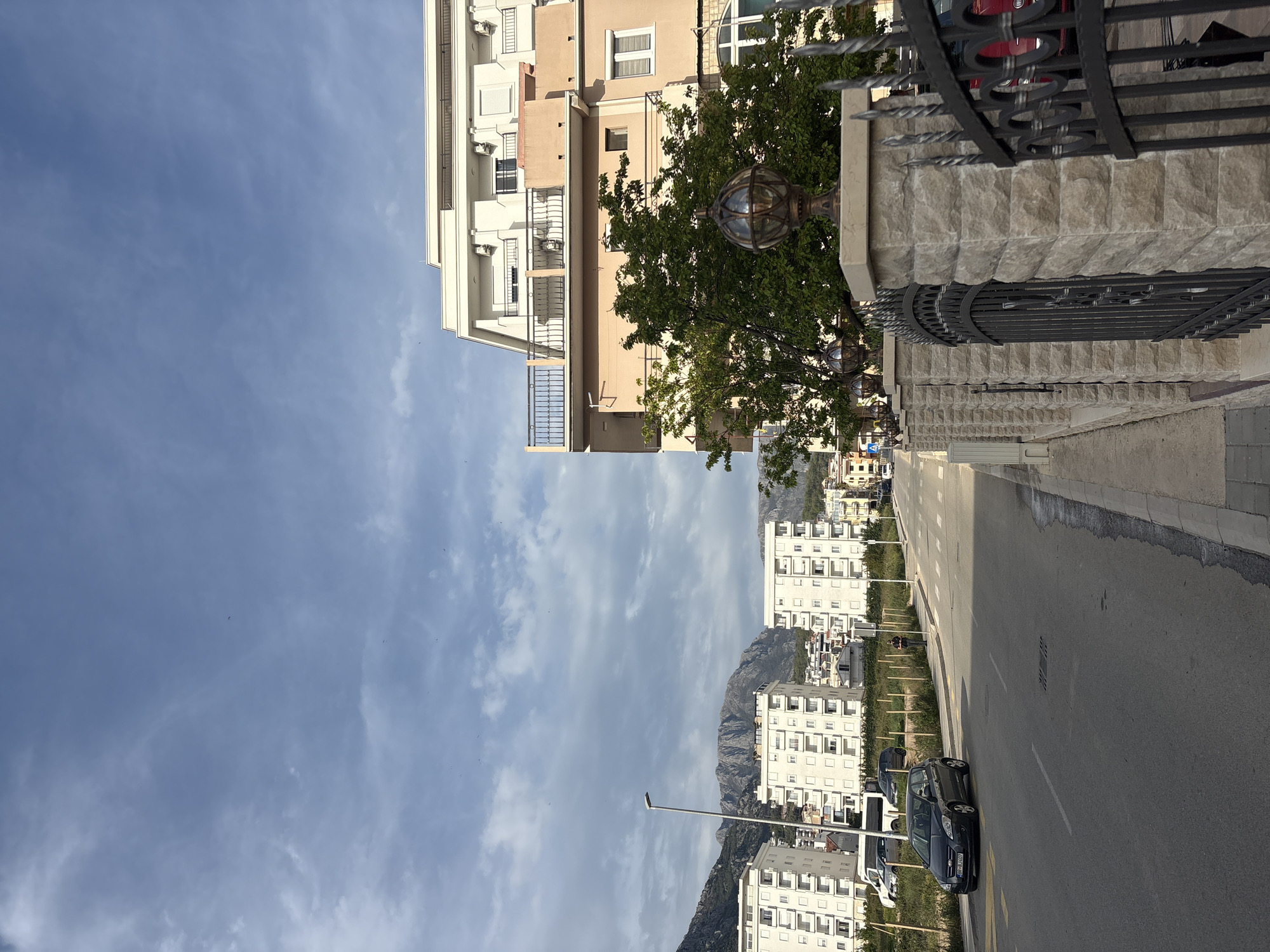 The image captures an upward-angled view of a street in Bar, Montenegro. A multi-story building, tan with white trim, stands on the left side. A wrought-iron fence and stone wall run along the right side of the street, partially obscured by a tree with decorative globe lights.
In the background, white residential buildings ascend toward a mountain range. The street has parked cars and streetlights. The sky is blue with scattered clouds.