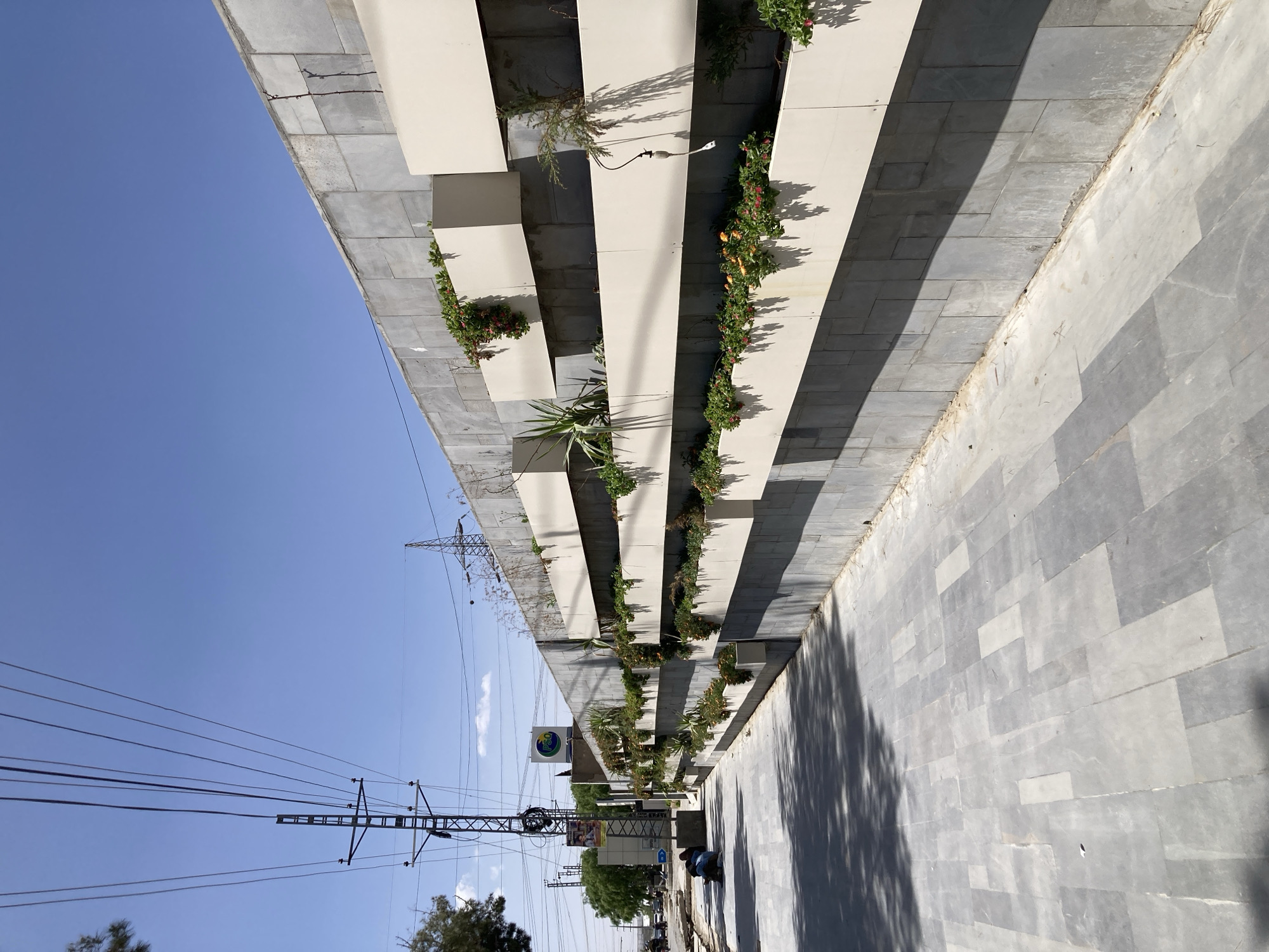 The image depicts an outdoor urban street scene in Quetta City Tehsil, Pakistan, under a bright, clear blue sky. On the left side, a modern building facade or wall features a series of stacked, light-colored, rectangular planter boxes arranged horizontally in a staggered, tiered pattern. These planters are filled with diverse greenery, including flowering plants with red and orange blossoms, succulents, and small shrubs, with some foliage trailing down the sides. Sunlight casts sharp, dark shadows from these structures and plants onto the grey, tiled wall behind them and the paved ground below.
The street and ground area on the right are covered with rectangular grey tiles of varying shades, some appearing lighter, and show distinct shadows, likely from trees or other overhead objects. In the distance, a few individuals are visible on the street, with one person possibly seated.
Tall utility poles and electricity pylons, supporting a dense network of black overhead power lines, are prominent on the left against the blue sky. A round blue and green sign with a leaf emblem is visible, displaying the word "GO" at the top and likely "GREEN" or a similar word at the bottom. Further down the street, other buildings, indistinct signs, and faint outlines of vehicles like cars and motorcycles can be seen. The strong shadows and clear sky indicate a sunny day, likely midday or early afternoon.