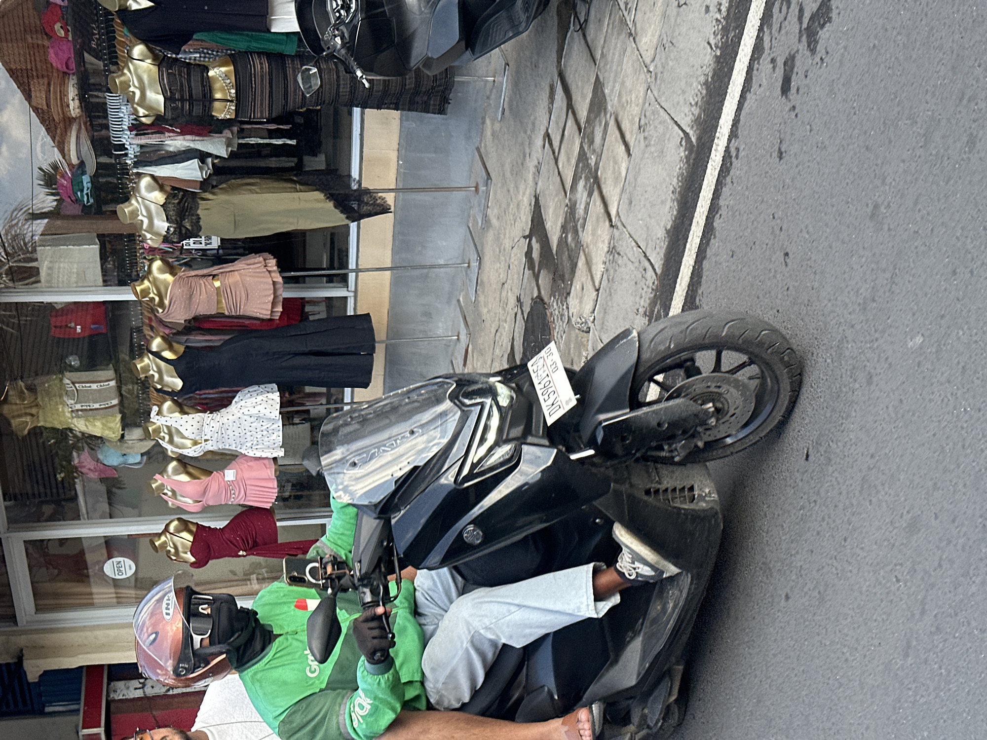 A person wearing a green jacket and a helmet is riding a black scooter on a street in North Kuta, Indonesia. The person is looking at their phone, which is mounted on the scooter's handlebars. In the background, there is a storefront with mannequins displaying various dresses. The street is paved with gray concrete tiles and has a white line marking the edge of the road. The scooter has a license plate with the text "DK 5961 FS" and "03-30". The weather appears to be overcast.