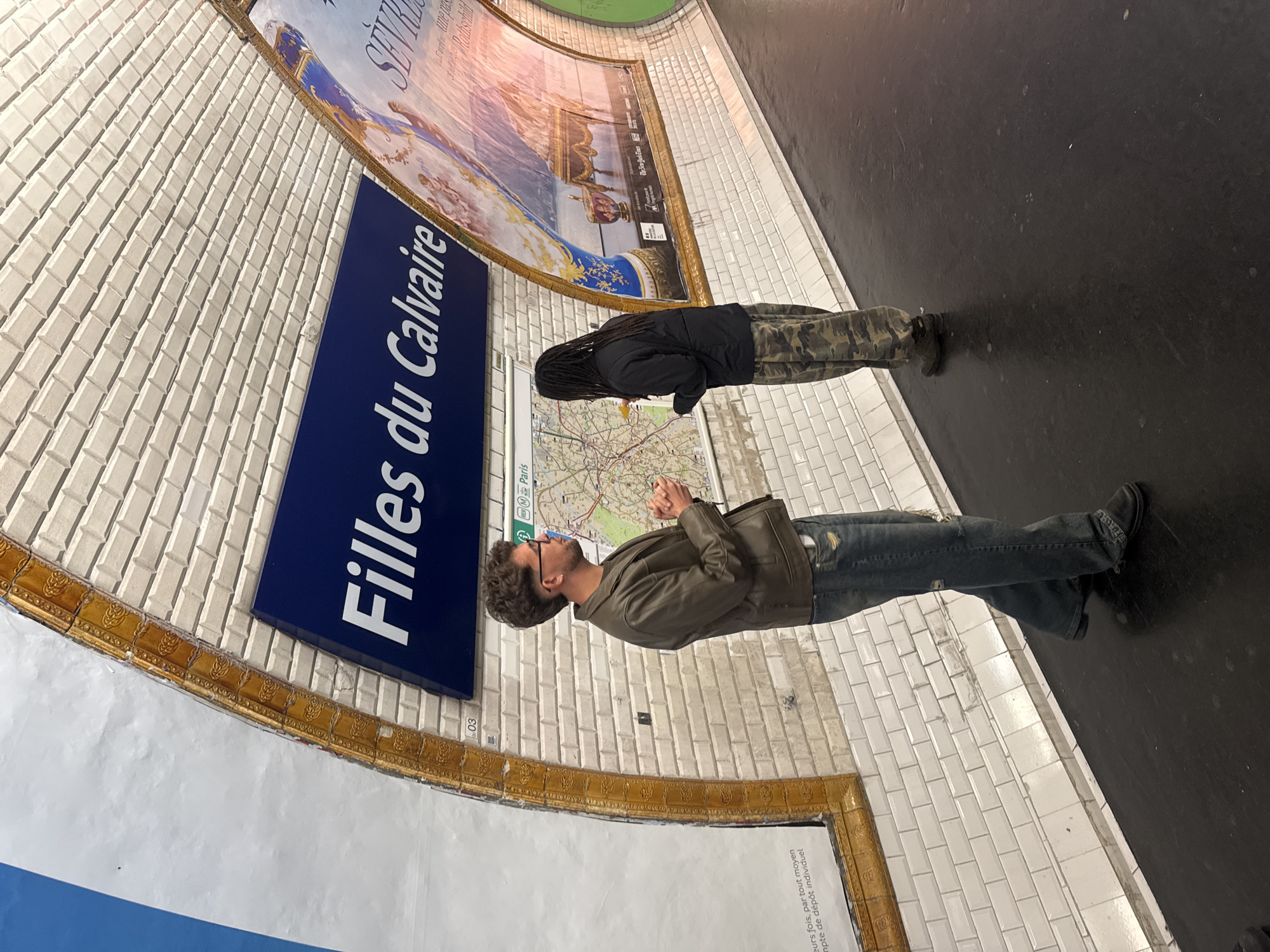 The image shows two individuals inside a Parisian metro station. The station is identifiable by the "Filles du Calvaire" sign. One person, wearing a brown jacket and jeans, is standing with their hands clasped, looking at a metro map. The other, clad in a black jacket and camouflage pants, is also looking at the metro map. The walls are tiled in a beige color, and there is a large advertisement above the station sign. The floor is black.