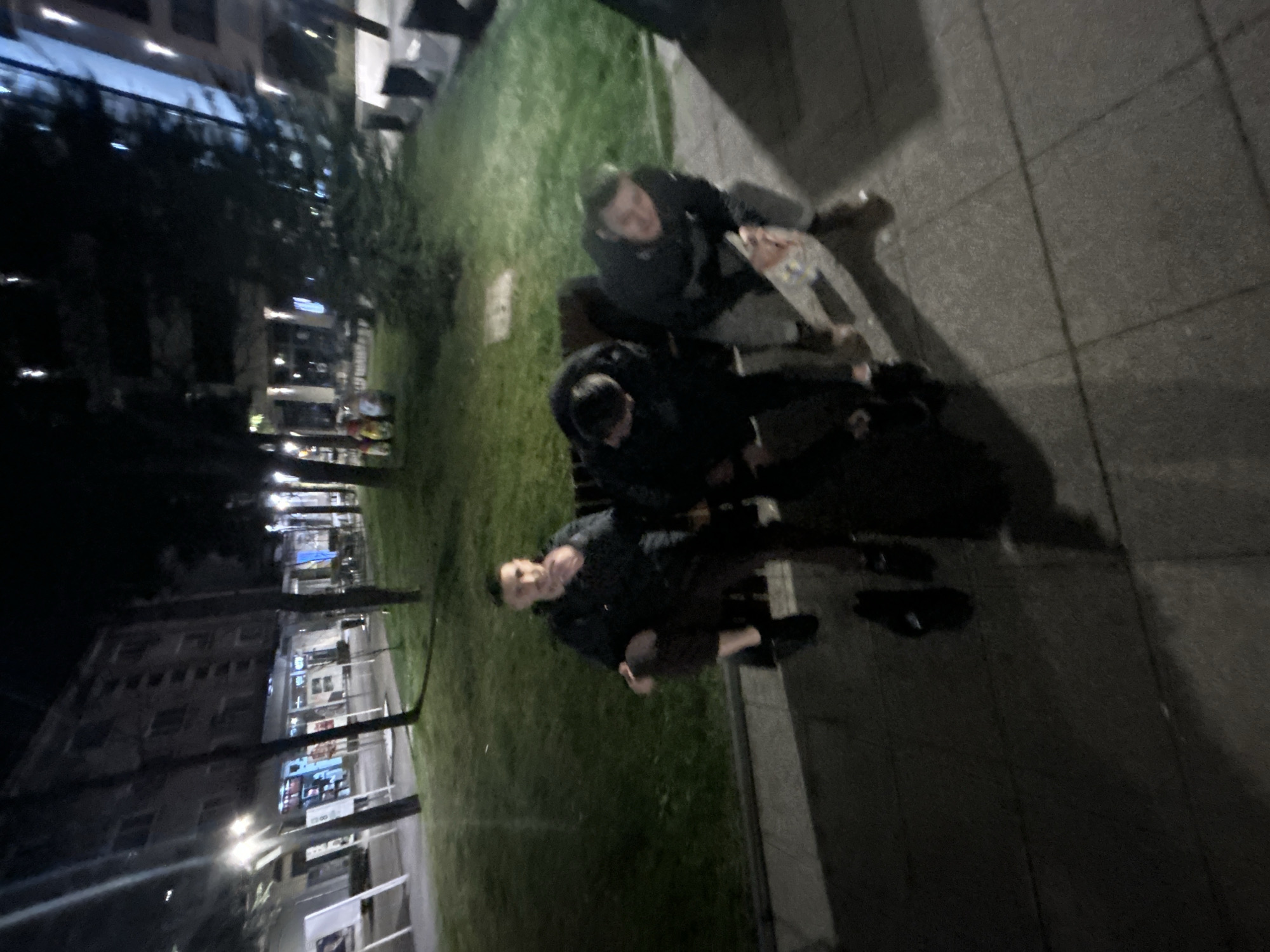 Night scene. Three people sitting outside in Vratsa, Bulgaria, near a building. Paved area and grass are visible.
