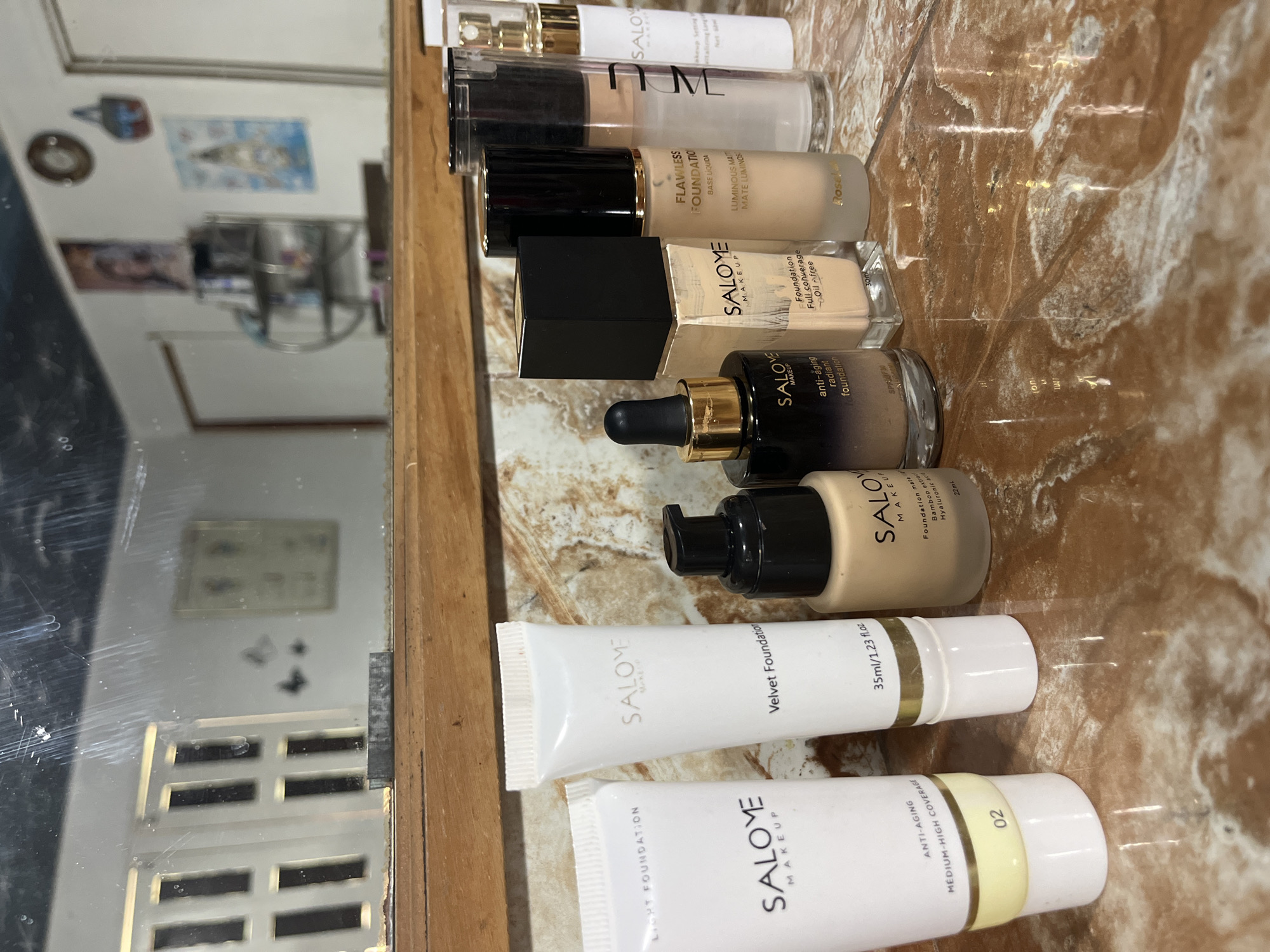 The image shows a collection of makeup products, primarily foundations, arranged on a reflective surface that appears to be a marble countertop. The products are mostly white tubes and glass bottles with black caps and gold accents. Some of the visible brands are "SALOME MAKEUP" and "ROSE GOLD." The items are positioned in a line, with some slightly overlapping. The background is a mirror reflecting a room with windows, shelves, and artwork on the walls. The lighting suggests an indoor setting, likely a home or a retail display. The city of Juan Griego, Venezuela, is not visually represented in the image; the scene is devoid of any explicit geographical markers or context linking it to that location.
