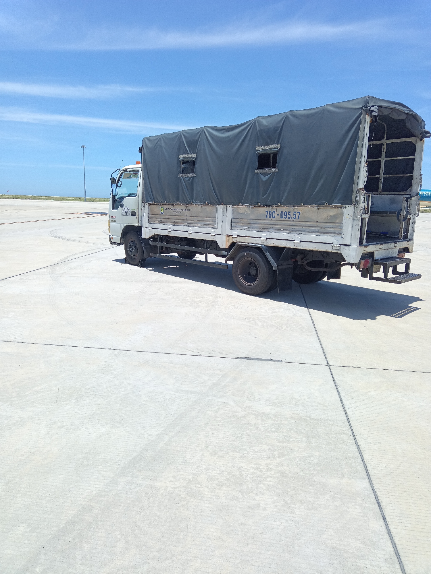 The image shows a white Isuzu truck parked on a concrete tarmac, likely at an airport or industrial area. The truck has a dark gray tarp covering its cargo bed, with two small rectangular openings visible on the side. The license plate reads "79C-095.57". The truck is white with some rust and wear. The tarmac is marked with lines and tire tracks. In the background, a light pole and a blue sky with white clouds are visible. The environment appears to be outdoors in daylight. The truck is stationary and there are no people visible in the image. The location is identified as Bắc Cam Ranh, Vietnam, which is likely an airport.