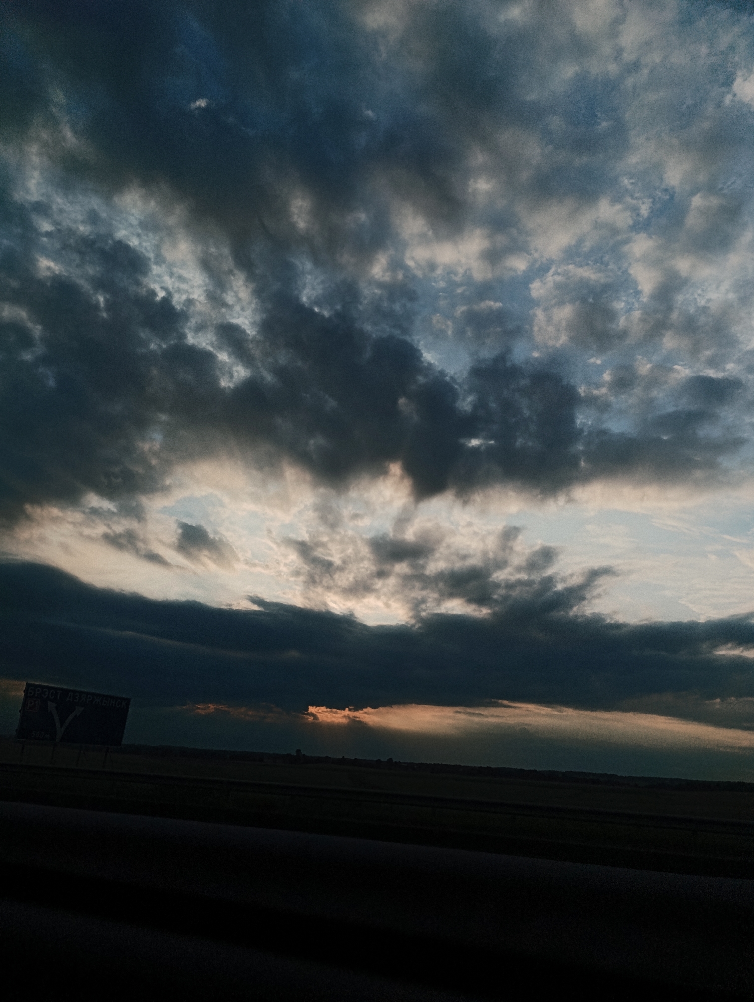 A wide-angle, low-angle shot shows a dramatic sky filled with dark, heavy clouds that obscure most of the sun. Sunbeams break through the clouds in the distance, casting a warm glow on the horizon. A dark, flat landscape stretches across the bottom of the frame, with a road or highway running through it. A dark sign with white text and a directional arrow is visible on the left side of the image, partially obscured by the low light. The overall mood is somber and foreboding, hinting at an impending storm.